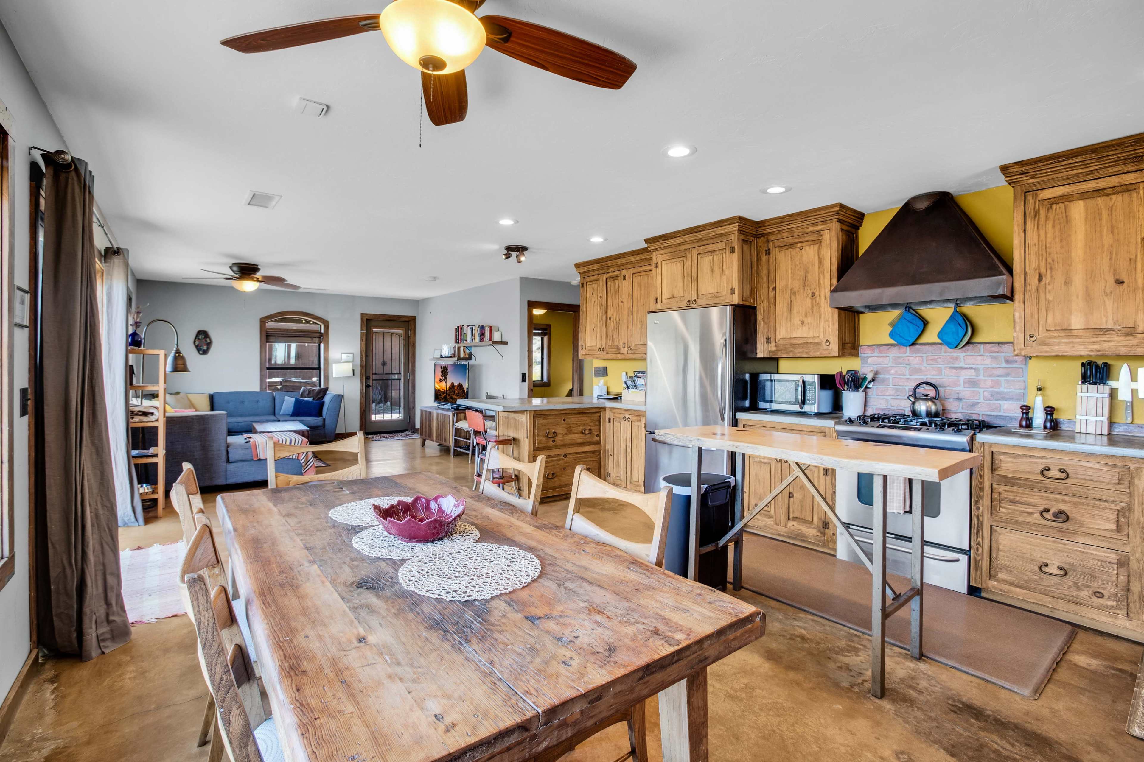 The image shows a modern kitchen with wooden cabinets and a large dining table, adjacent to a living area with a blue couch visible in the background.