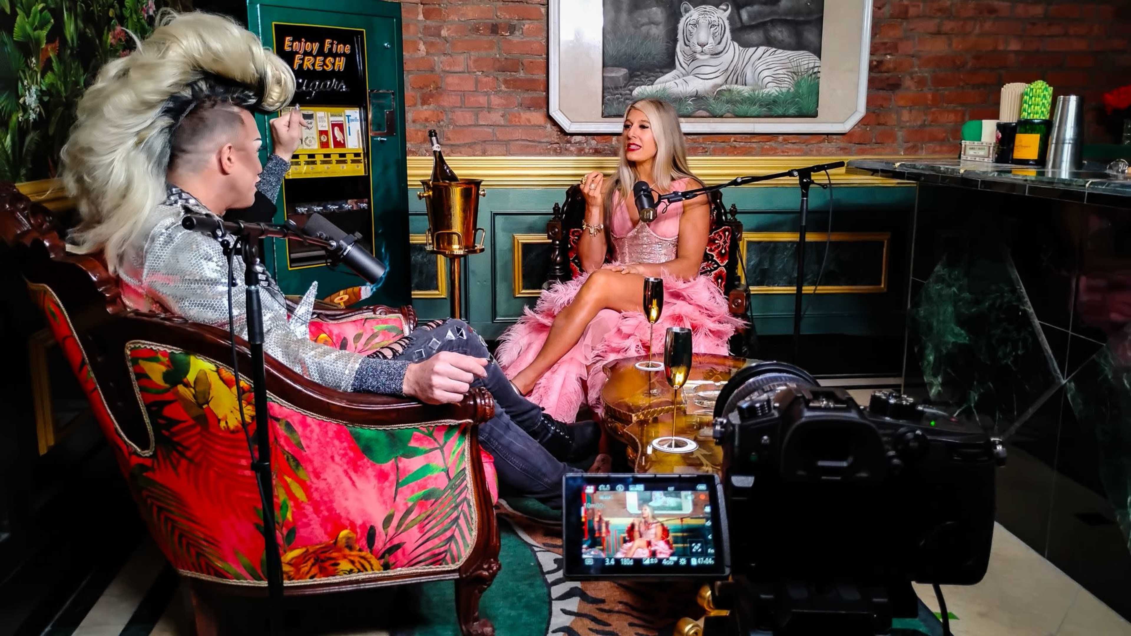 A man with a large hairstyle and a woman in a pink gown are seated in a decorated room, engaged in conversation while a camera captures the scene.