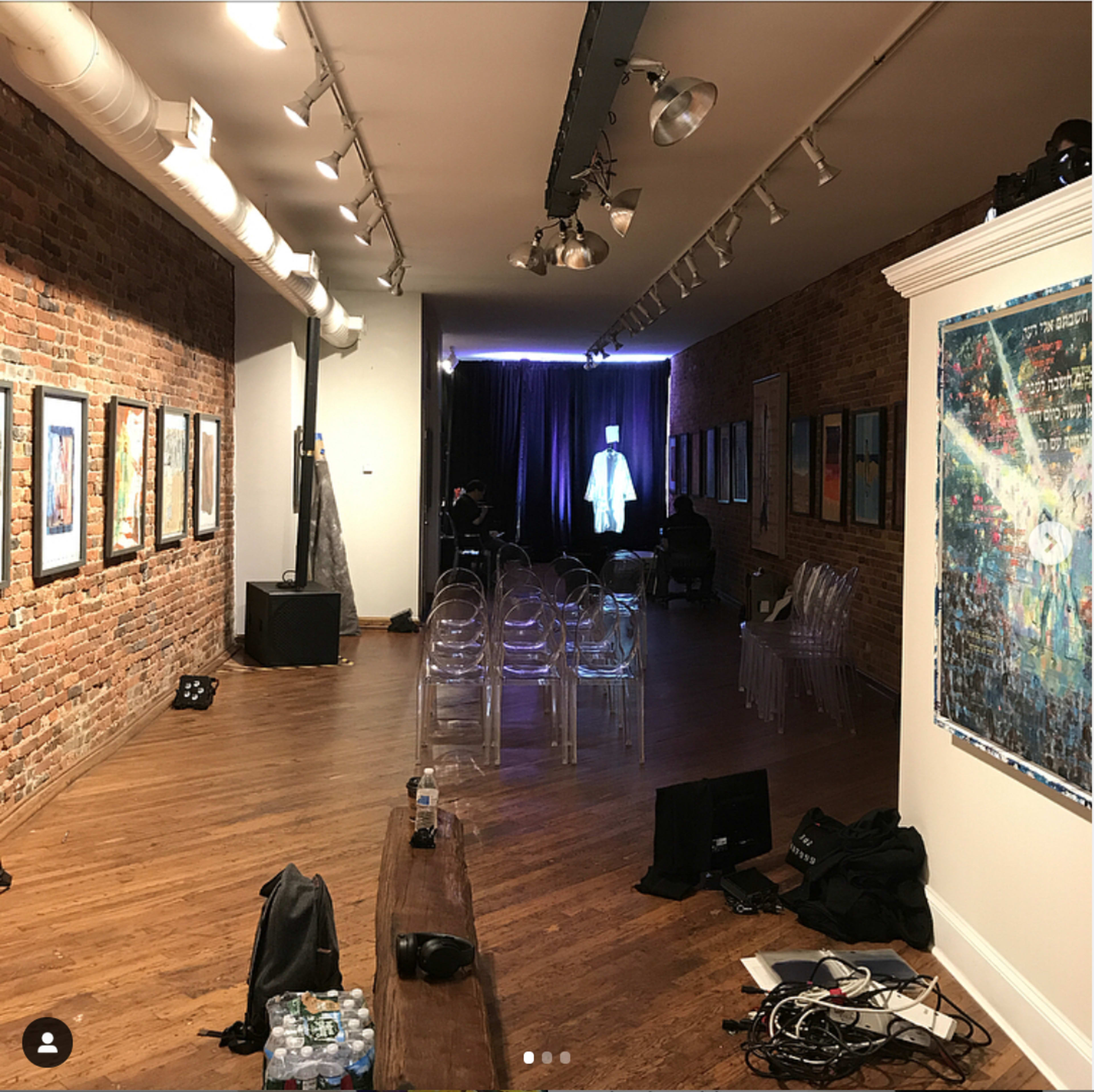 An art gallery with exposed brick walls, featuring framed artwork, transparent chairs arranged for seating, and a stage area with a curtain in the background.