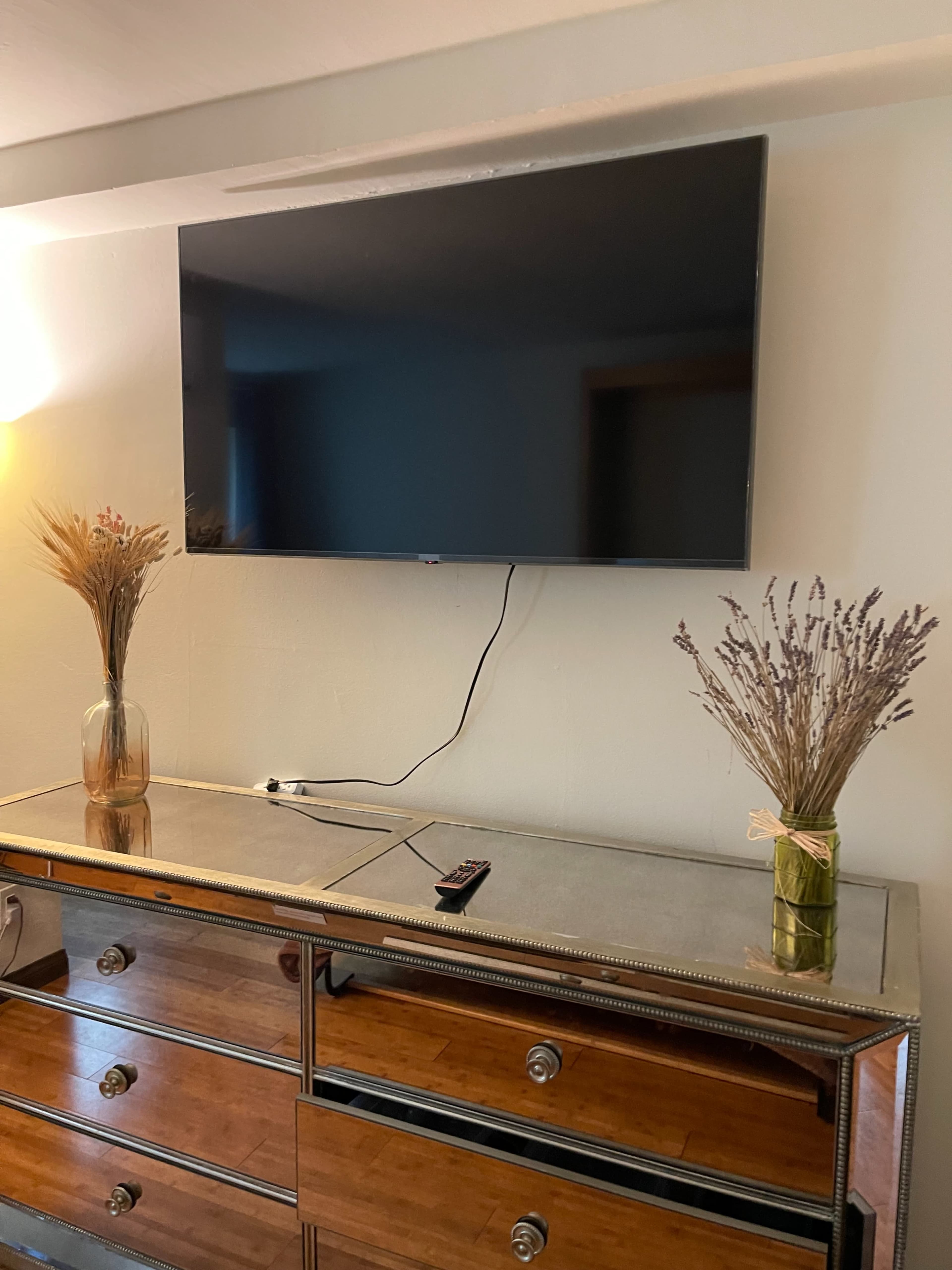 A flat-screen TV is mounted on a wall above a decorative dresser, with two vases of dried flowers on the dresser and a remote control lying on its surface.