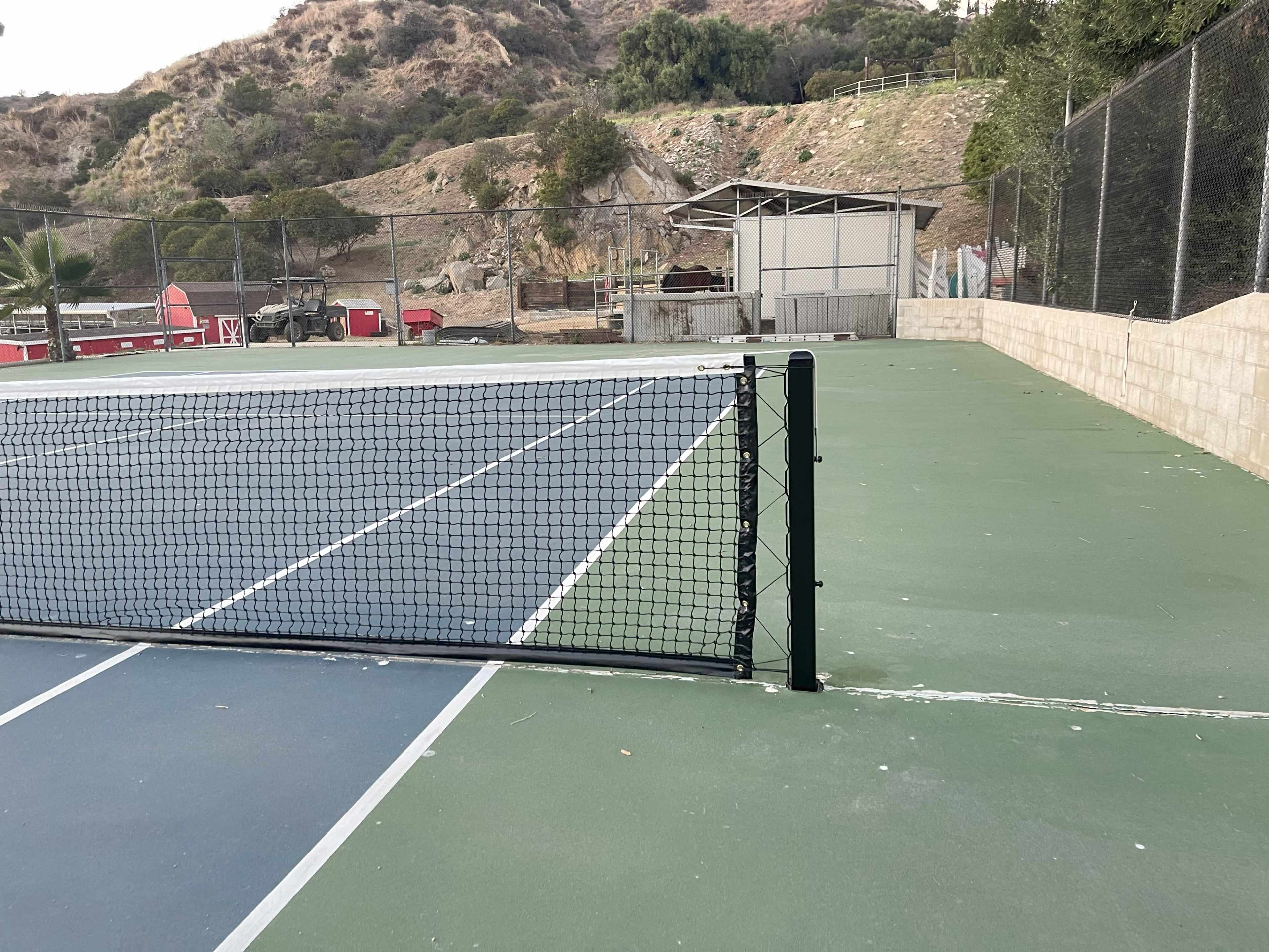 A tennis court with a net in the foreground is situated near a hillside, with a storage shed and scattered vegetation in the background.