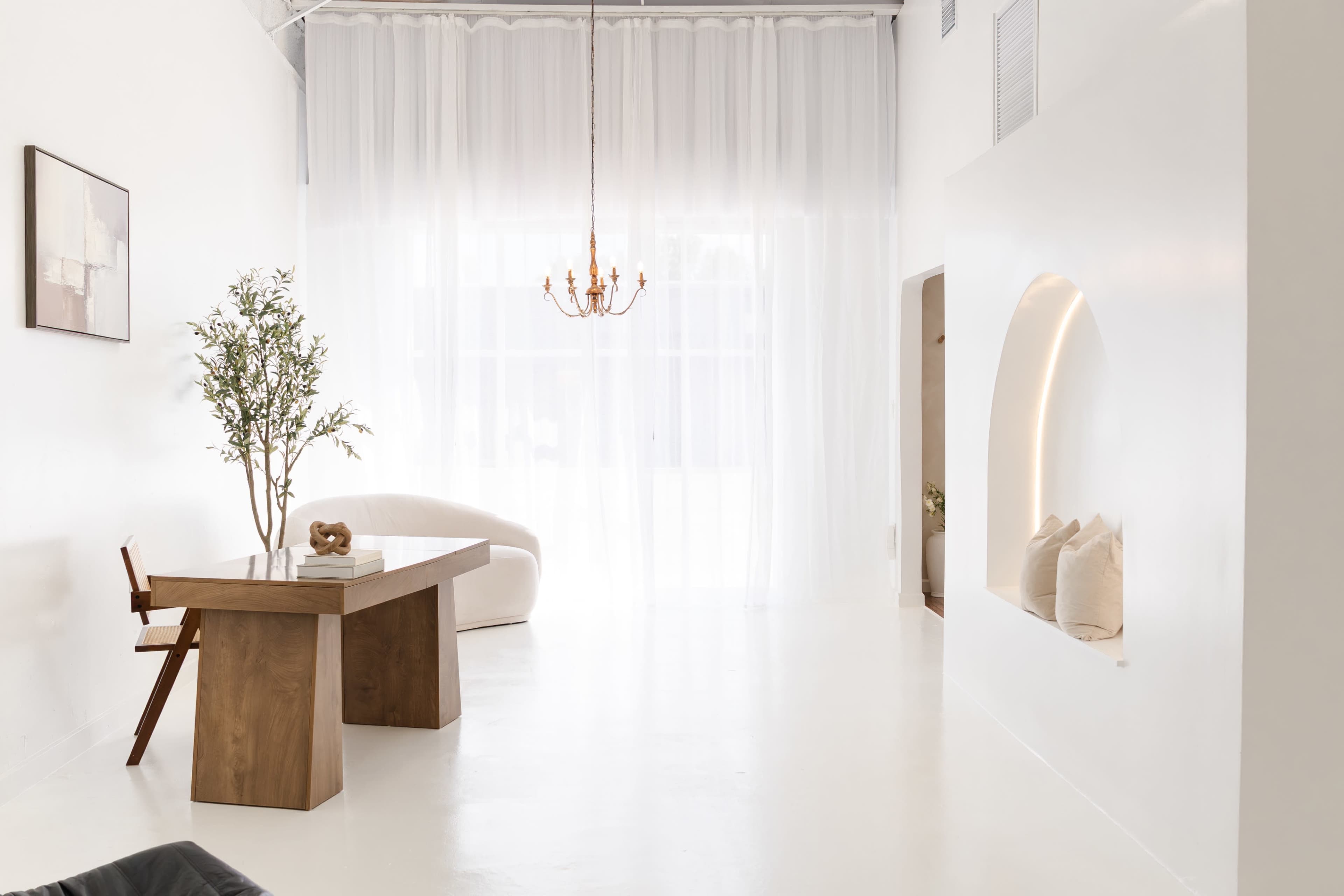 The image shows a modern, minimalist interior with a wooden dining table, a curved white sofa, a chandelier, and large sheer curtains filtering natural light.