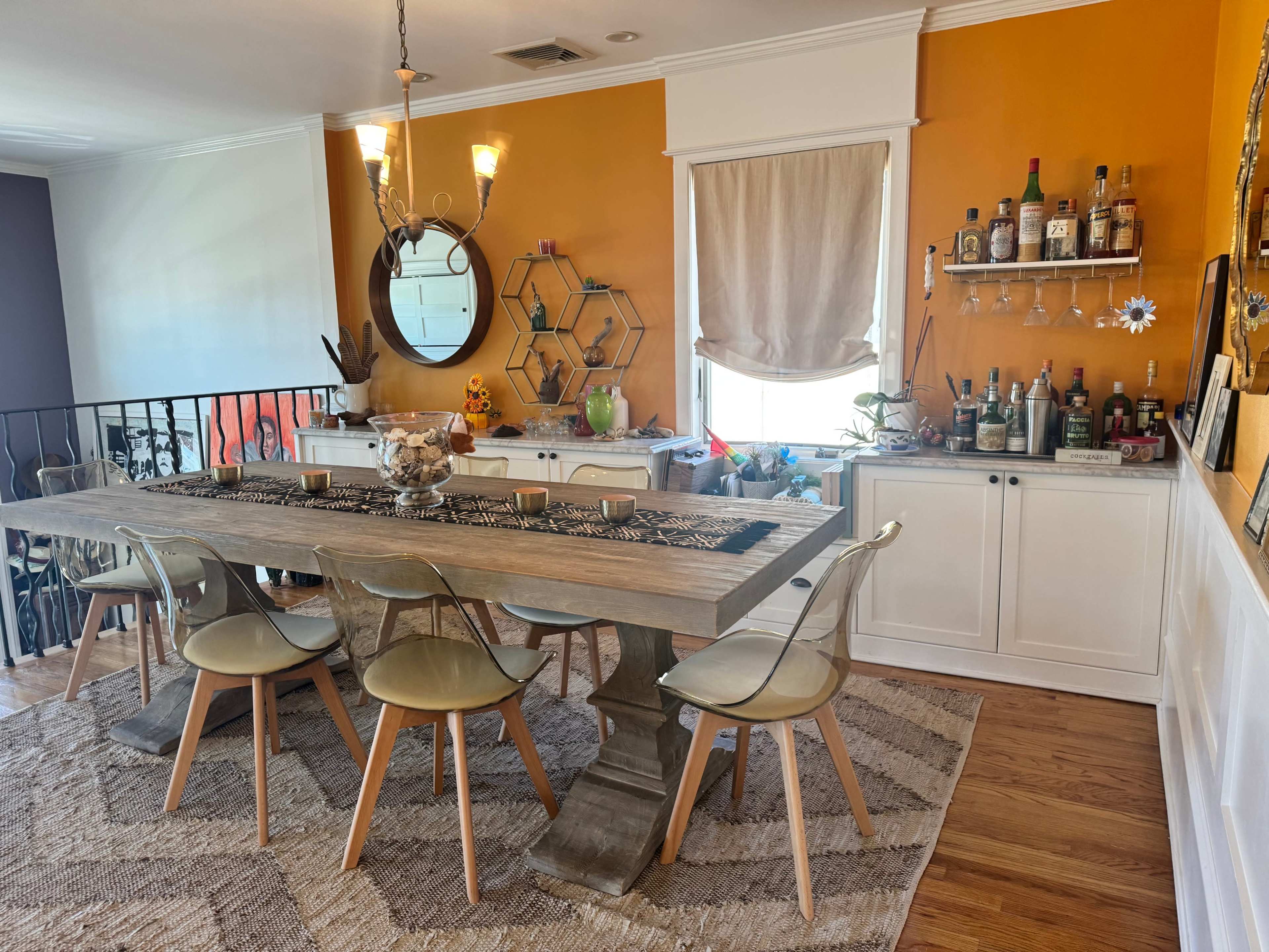 A modern dining area features a long wooden table surrounded by transparent chairs, with a bar cabinet displaying various bottles against a vibrant orange wall.