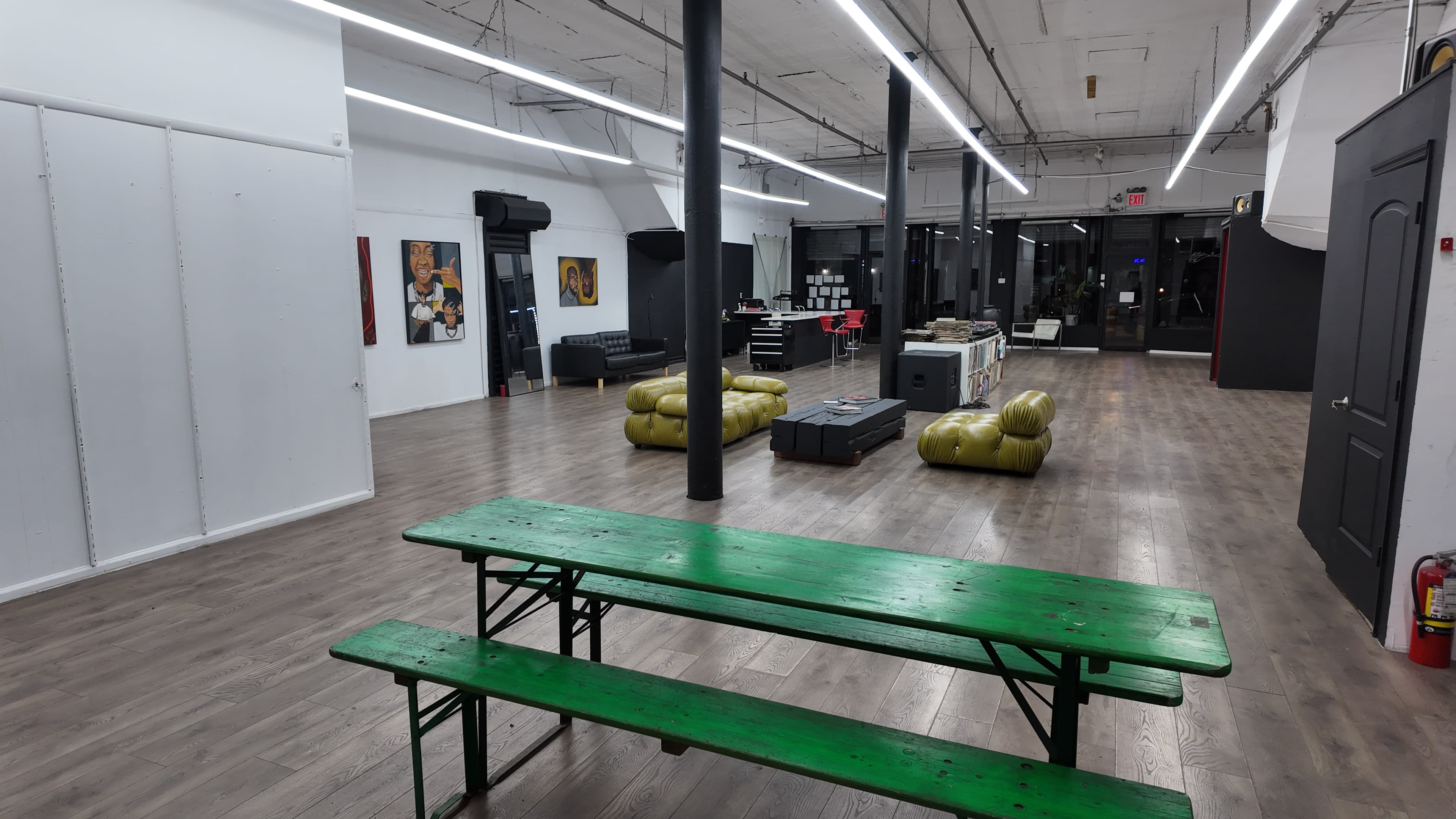 The image shows a spacious, empty interior of a modern commercial space with wooden floors, bright overhead lights, a green picnic table, and a few minimalist furniture pieces.