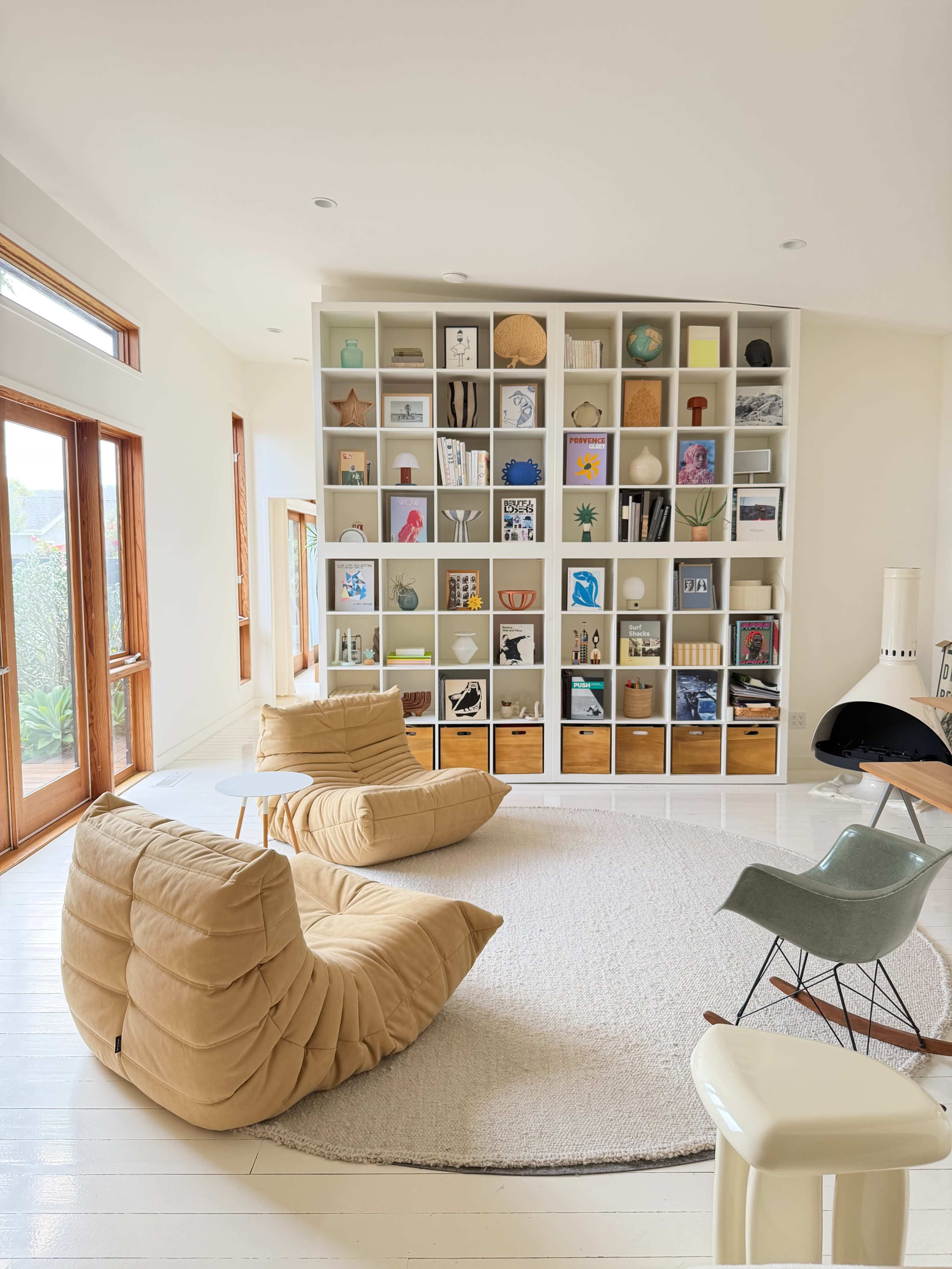 The image shows a modern living room featuring a large bookshelf filled with various decorative items and books, two padded chairs, a round rug, and large windows.