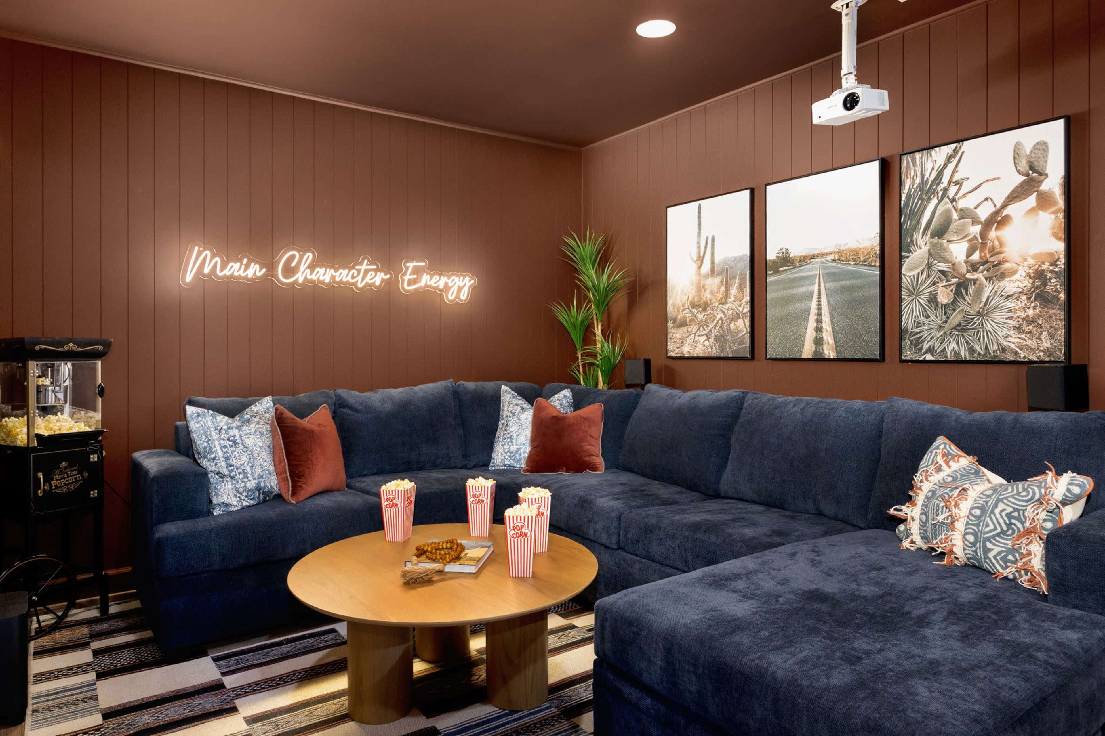 The room features a large blue sectional sofa, a wooden coffee table, and three framed pictures on the wall, alongside a neon sign reading "Main Character Energy" and a popcorn machine in the corner.