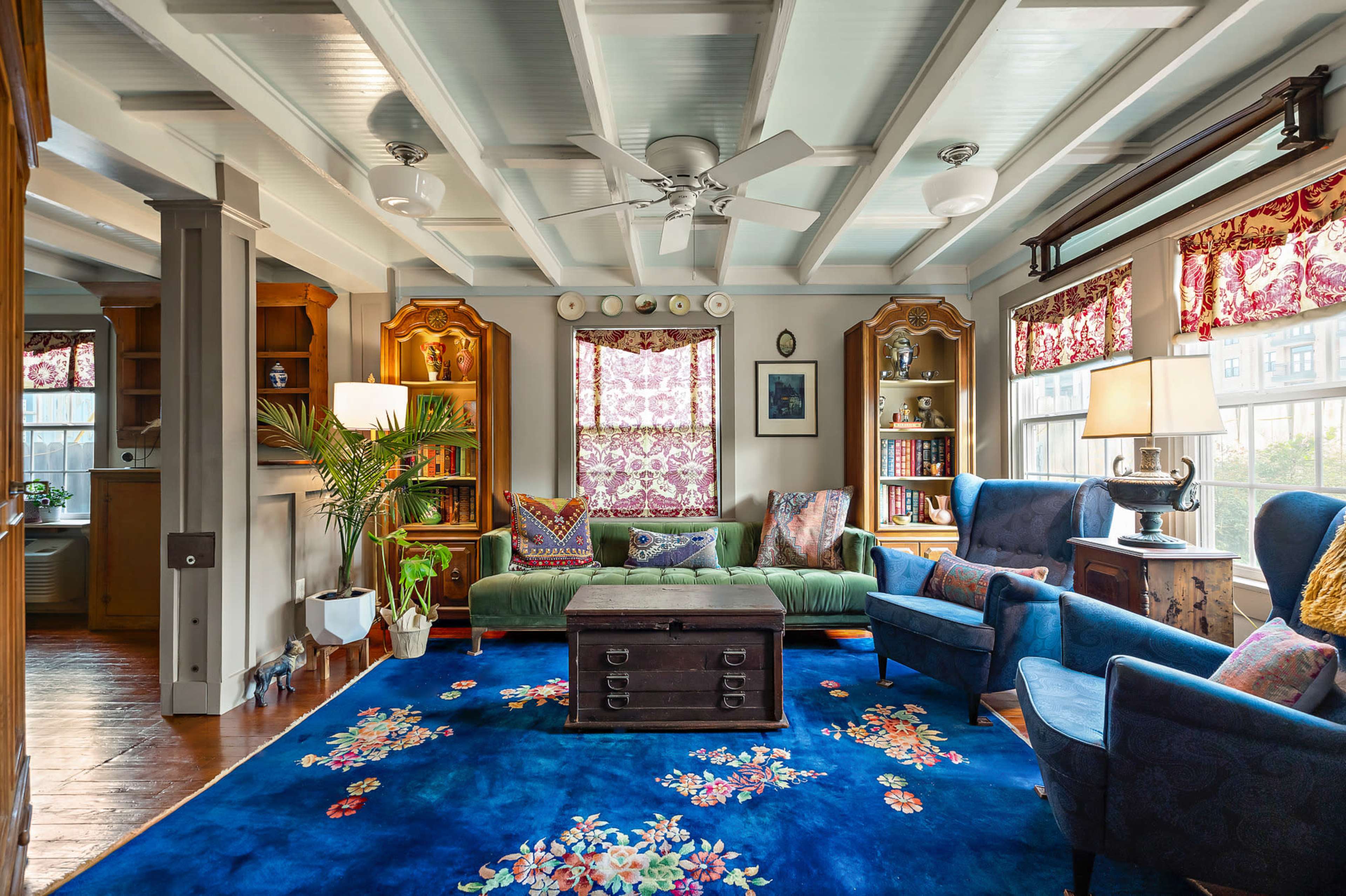 The image shows a cozy living room with a green sofa, two blue chairs, a dark wooden coffee table, and bookshelves filled with books and decorative items.