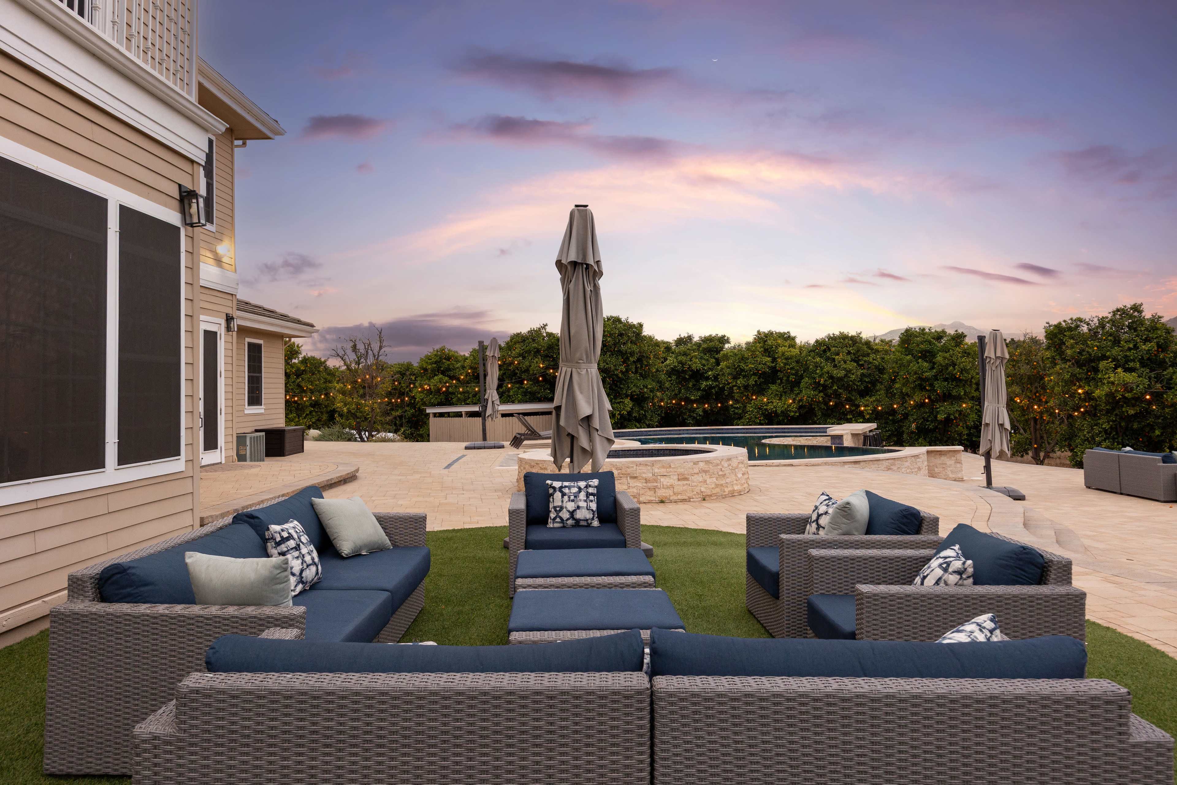 The image features a patio area with several outdoor couches arranged around a central space, alongside a pool, surrounded by carefully maintained landscaping under a colorful sunset sky.