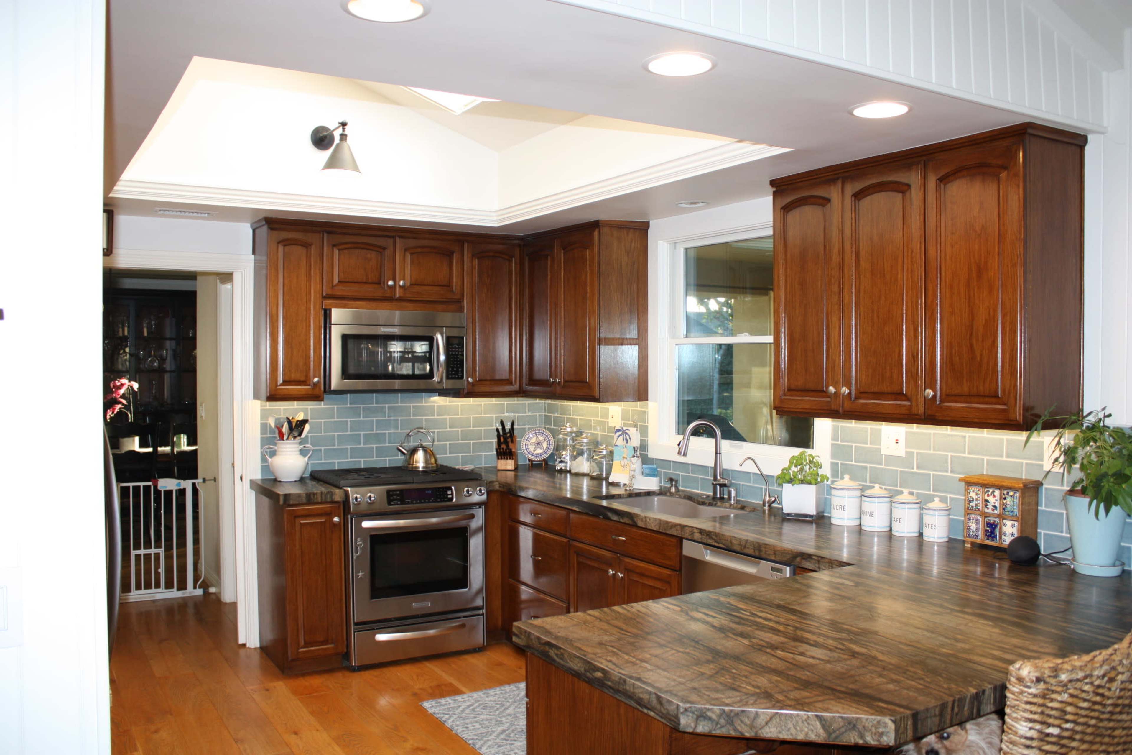 The image shows a kitchen with wooden cabinetry, a countertop, stainless steel appliances, and blue-tiled backsplash.