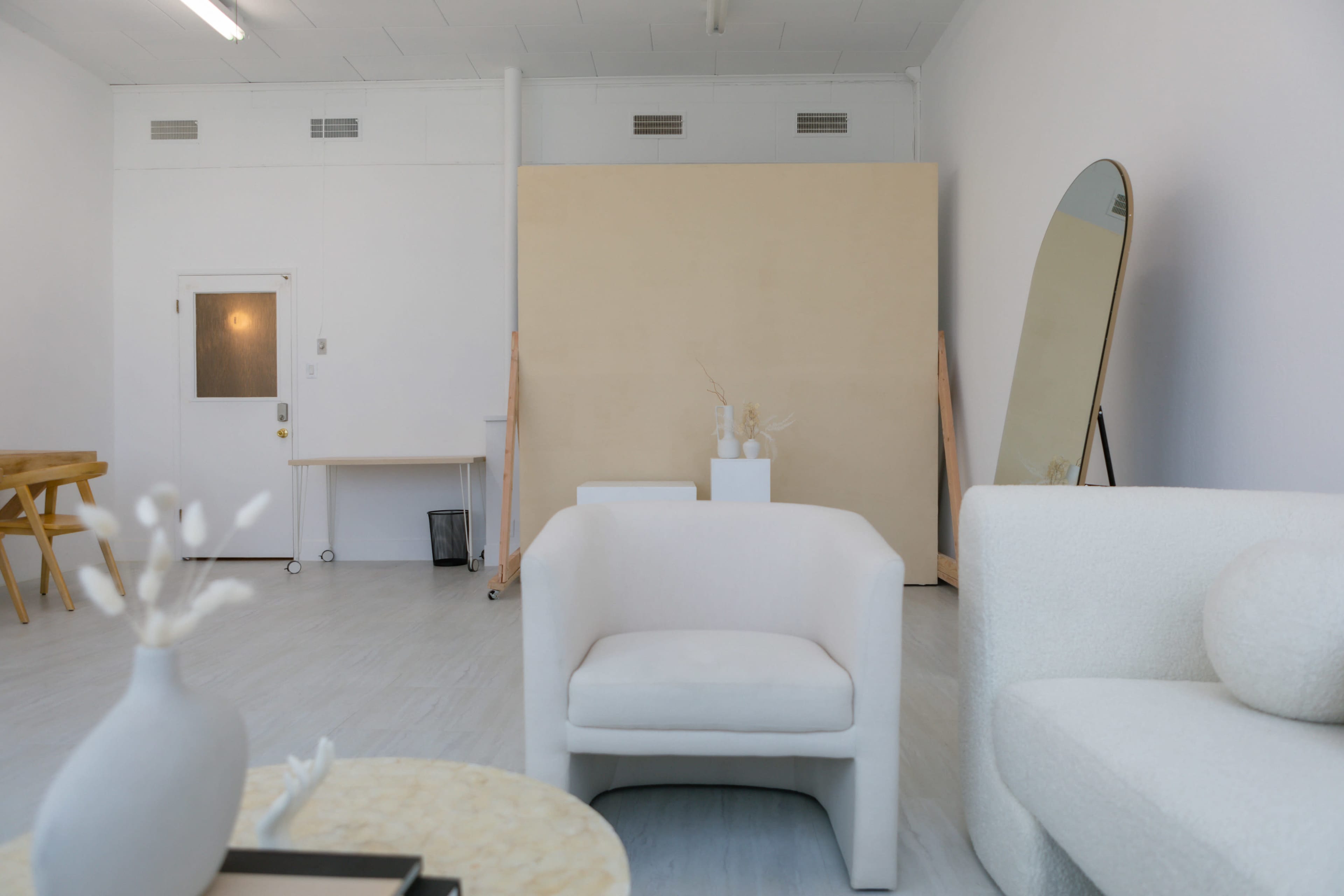 The image shows a spacious, minimalist studio with white walls, light wooden furniture, and a large mirror.