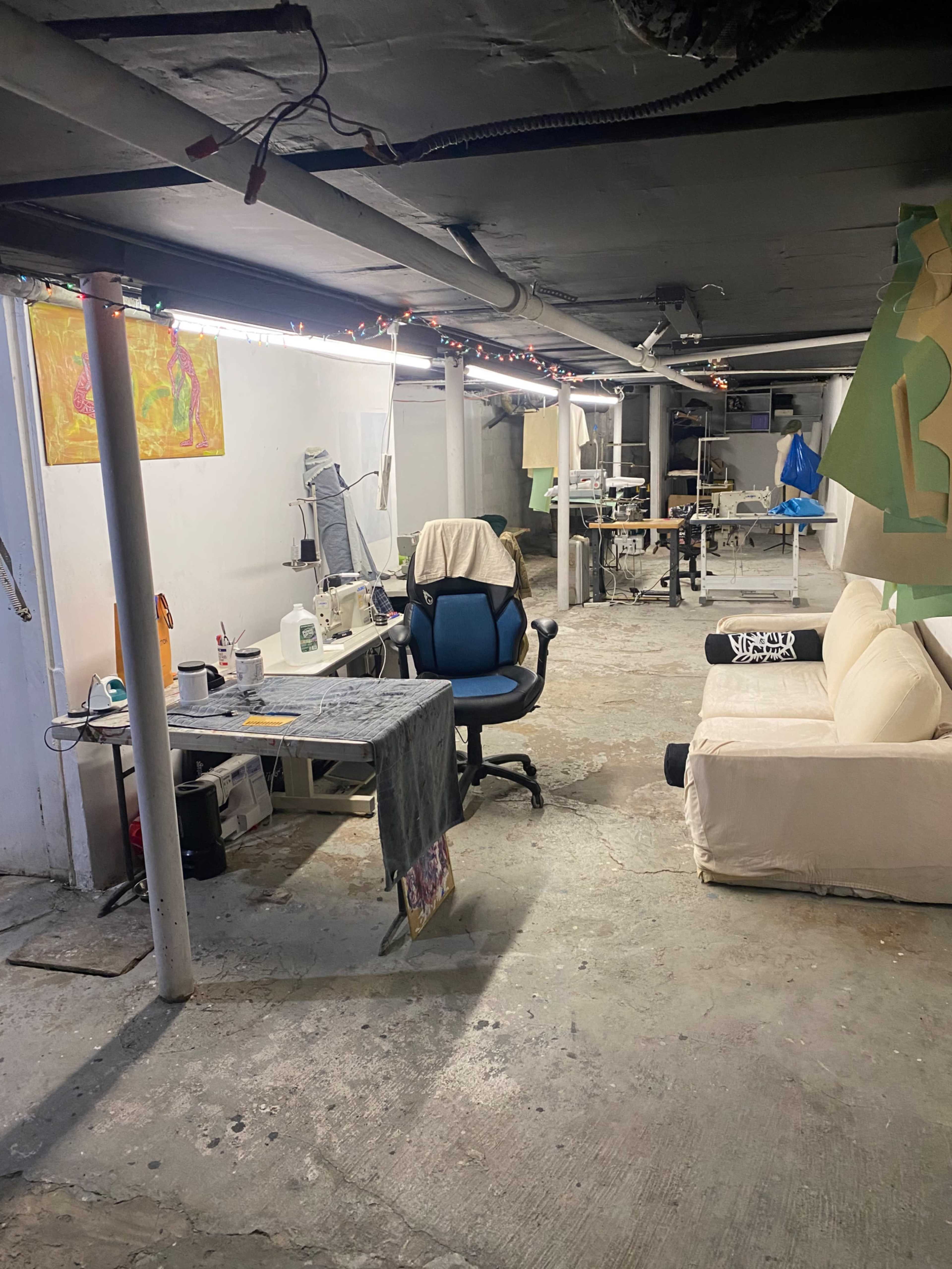 The image shows a spacious basement area with a sewing station, a blue office chair, a beige couch, and various art supplies scattered around.
