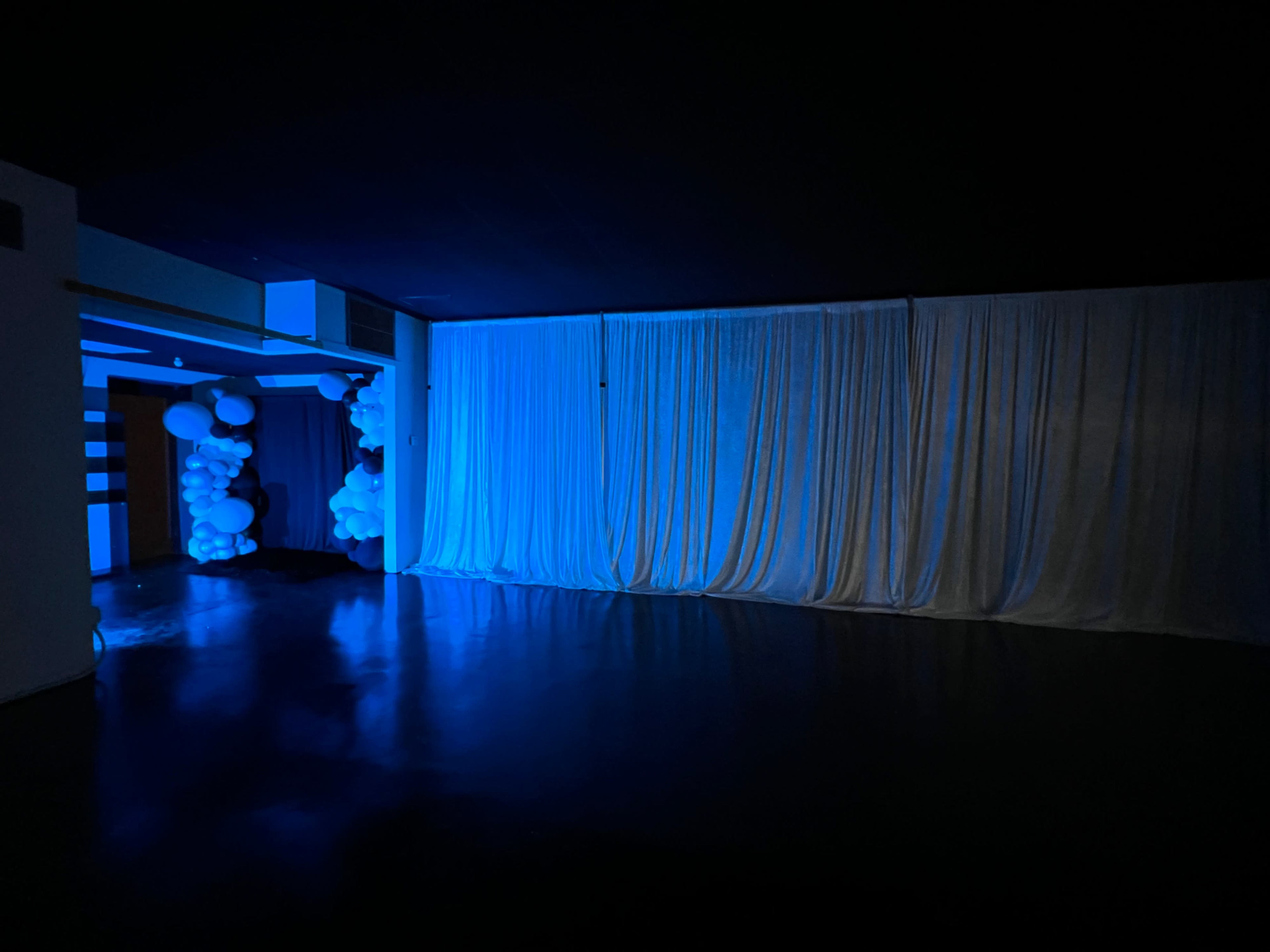 A dimly lit room with blue lighting, featuring a white curtain covering a wall and balloon decorations in the corner.
