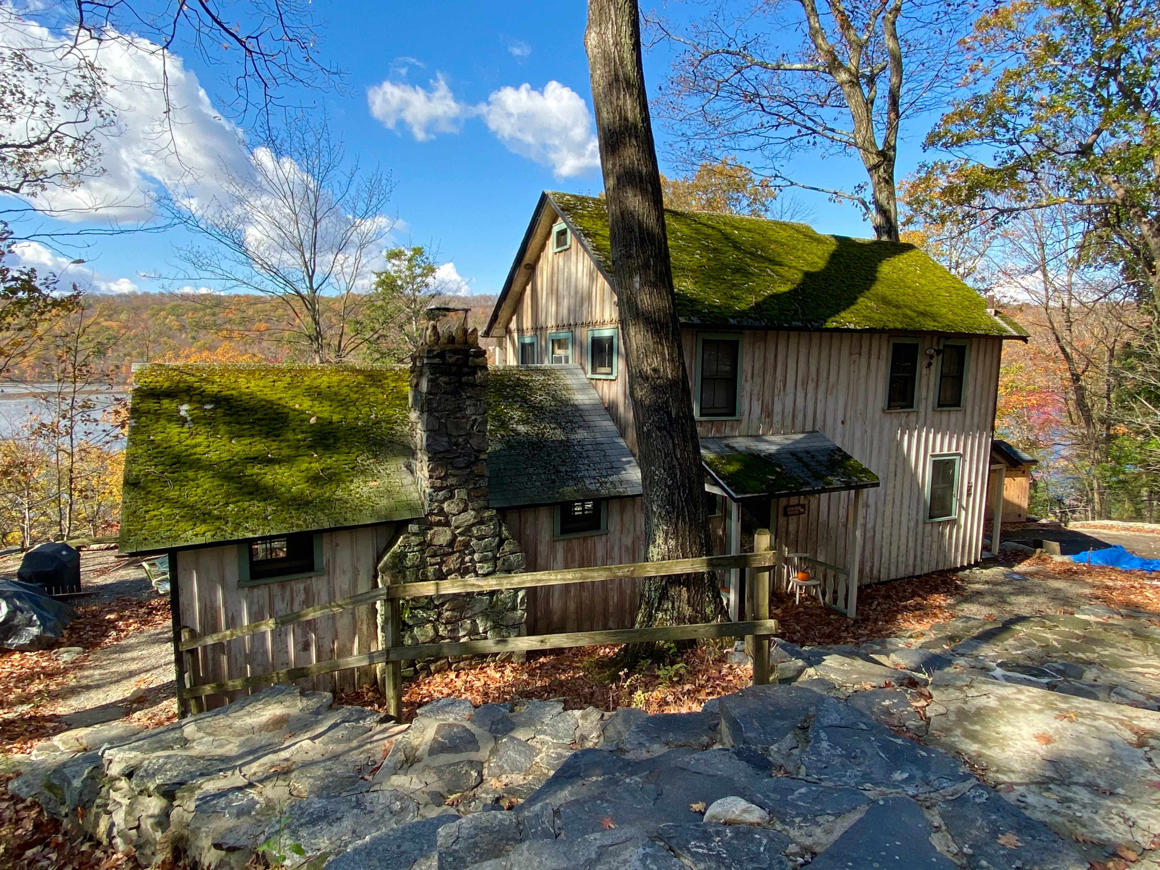 A wooden house with a moss-covered roof stands among autumn foliage near a lake.
