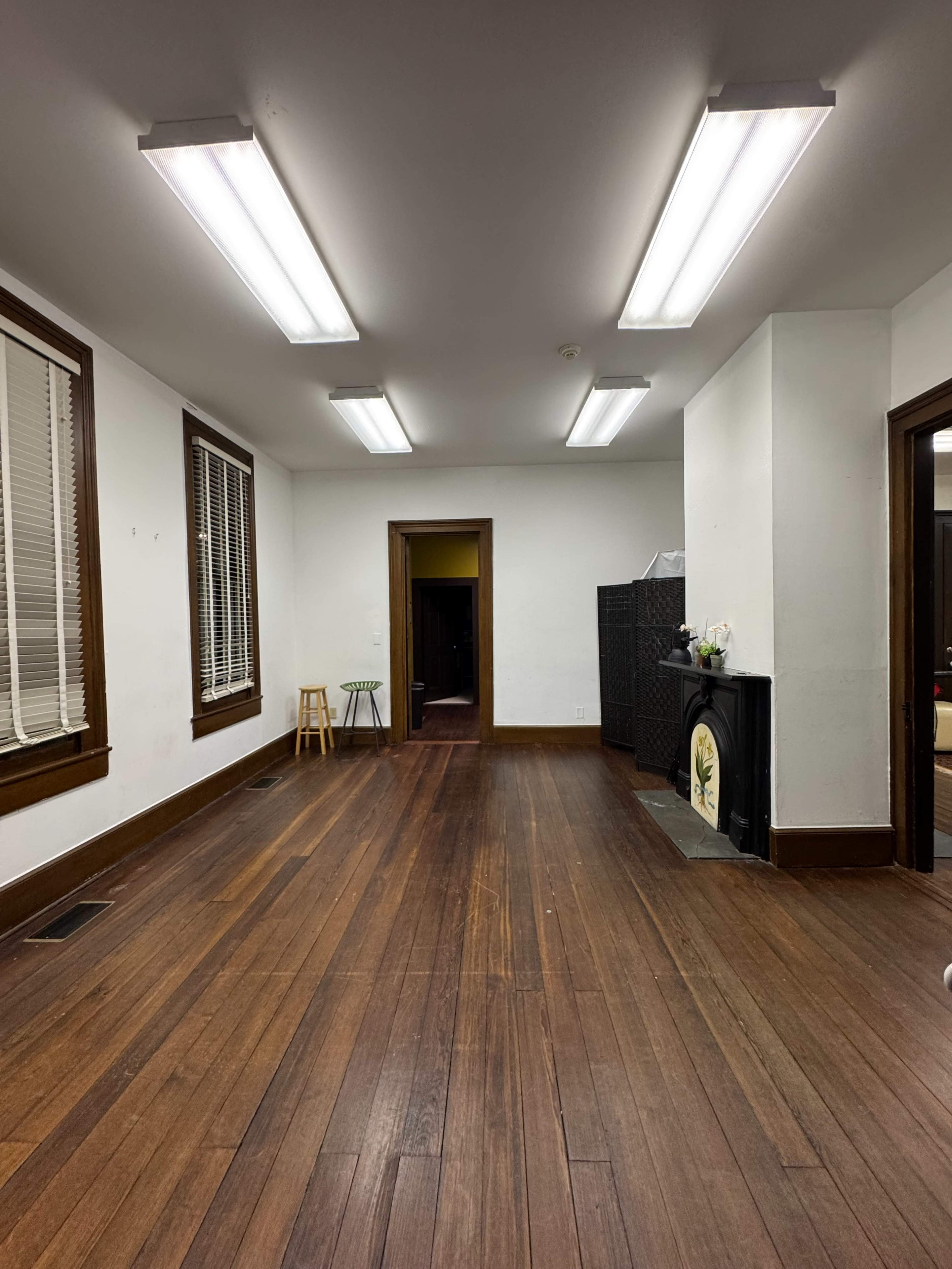 The image shows an empty room with wooden floors, white walls, fluorescent ceiling lights, and doors leading to other areas.