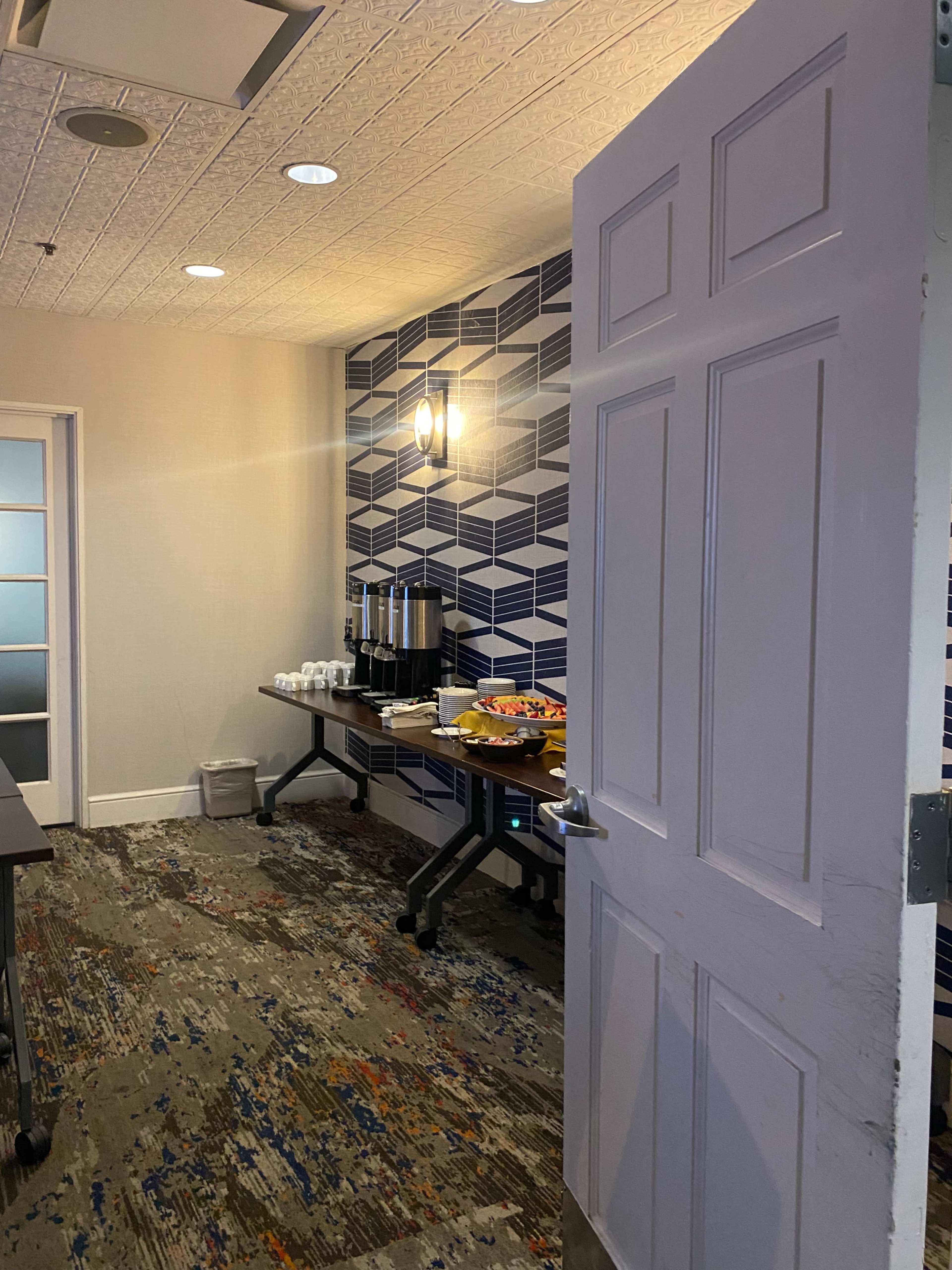 A doorway opens to a kitchenette area featuring a coffee station and refreshments on a table, set against a geometric-patterned wall.