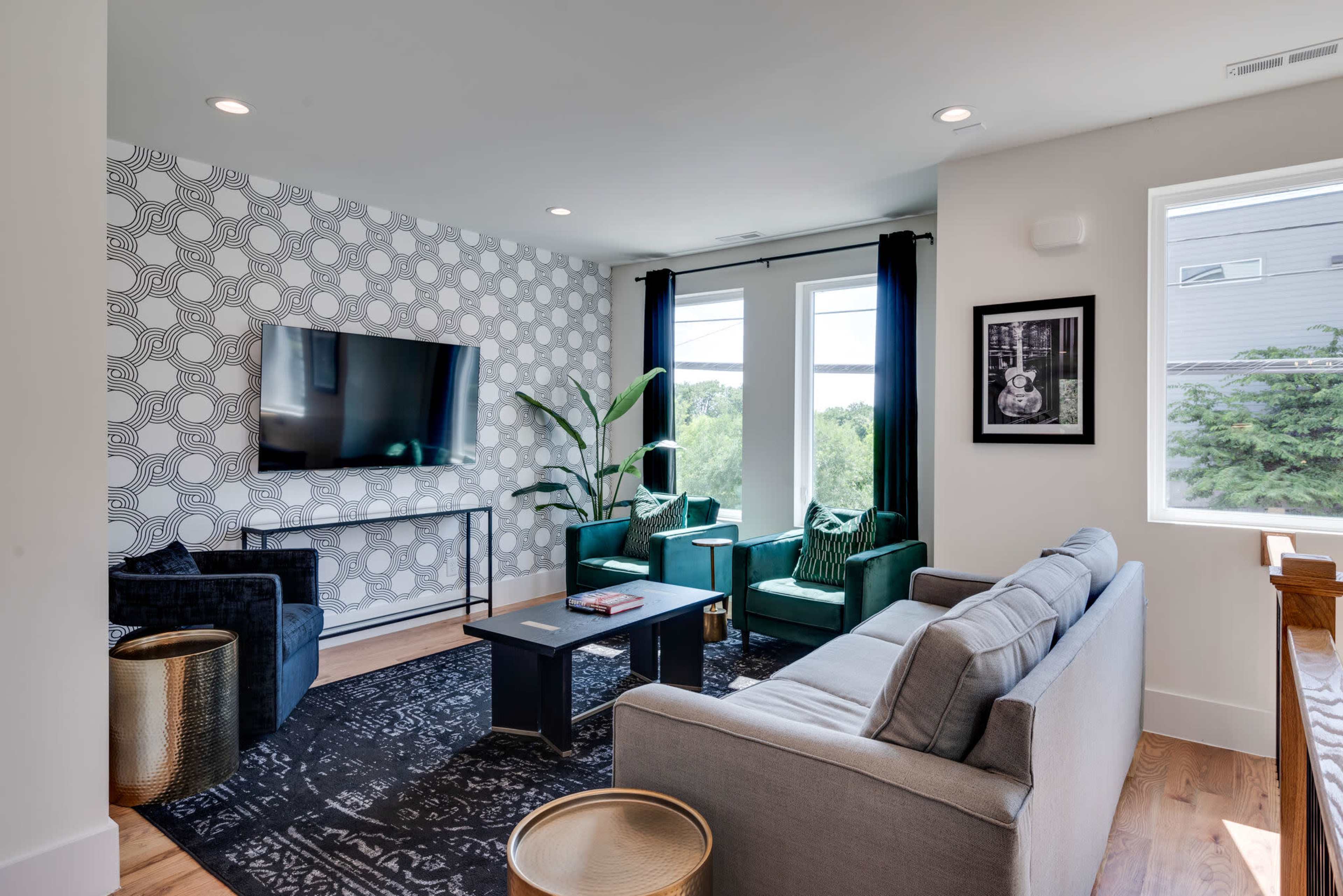 The image shows a modern living room with a patterned wallpaper, a large television on the wall, two green armchairs, a gray sofa, and a coffee table, all illuminated by natural light from large windows.