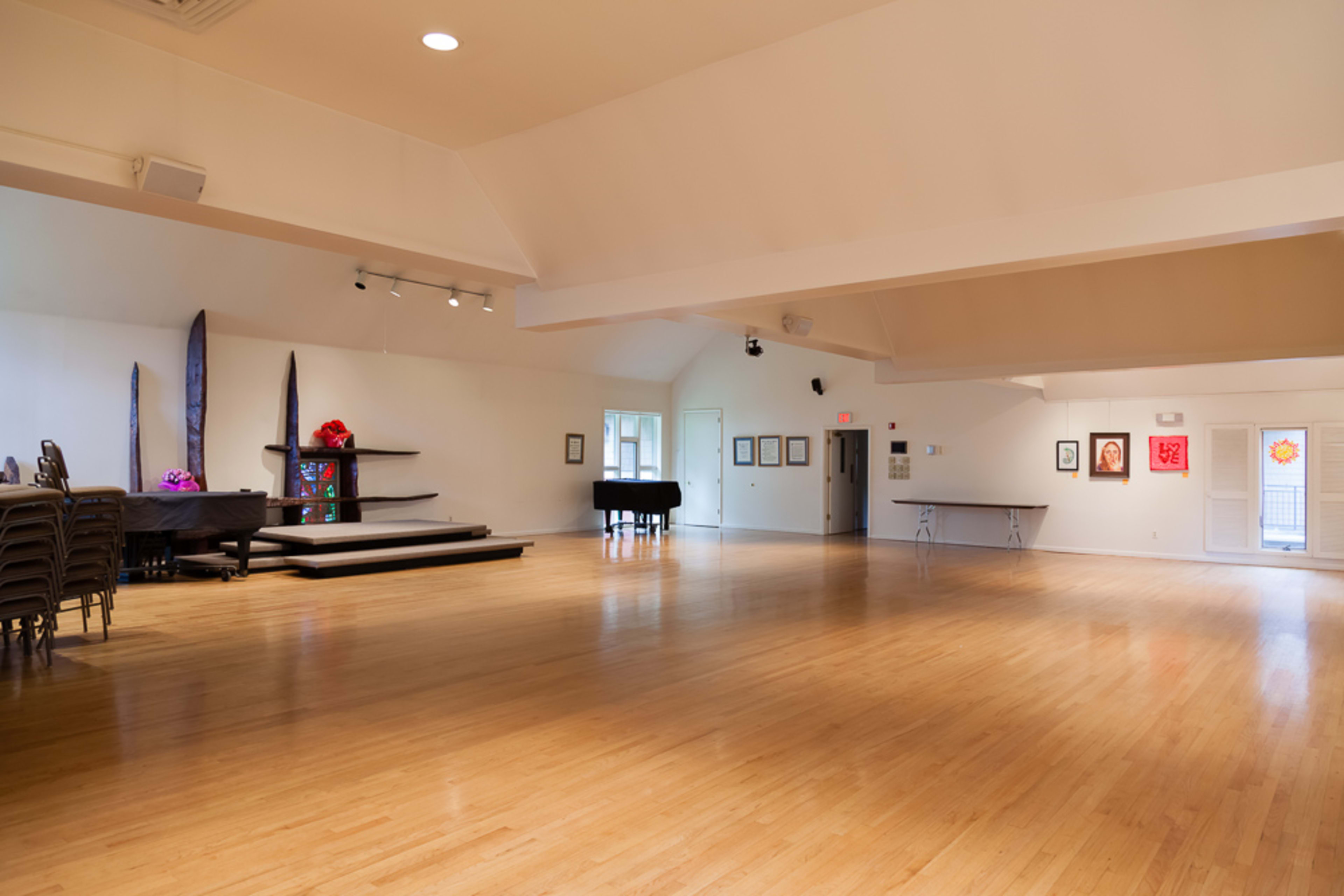 The image shows an empty multi-purpose room with wooden flooring, a stage, and several chairs stacked on the left side.