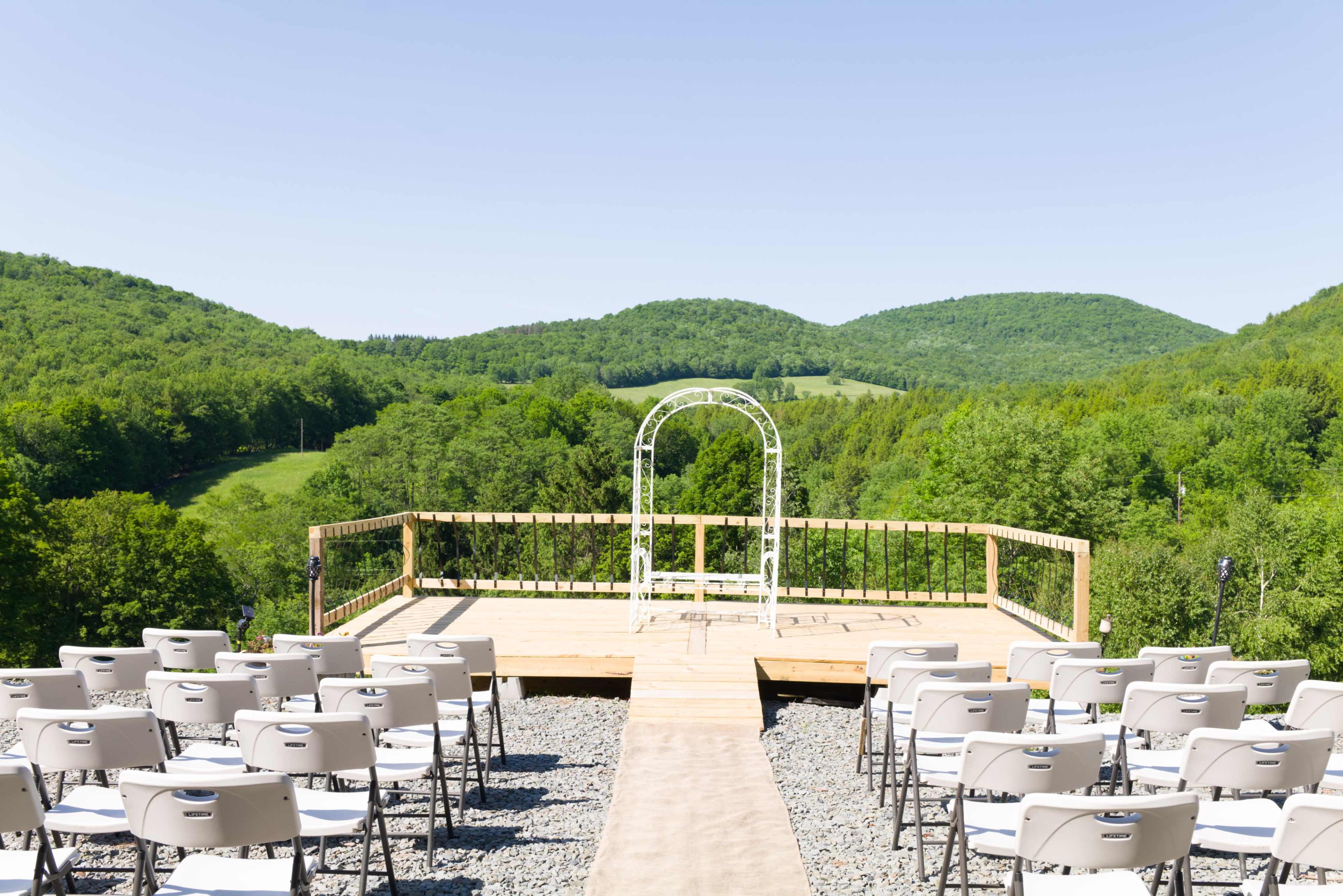 A wooden wedding altar is set up on a platform overlooking a green mountainous landscape, with rows of white chairs facing the altar.