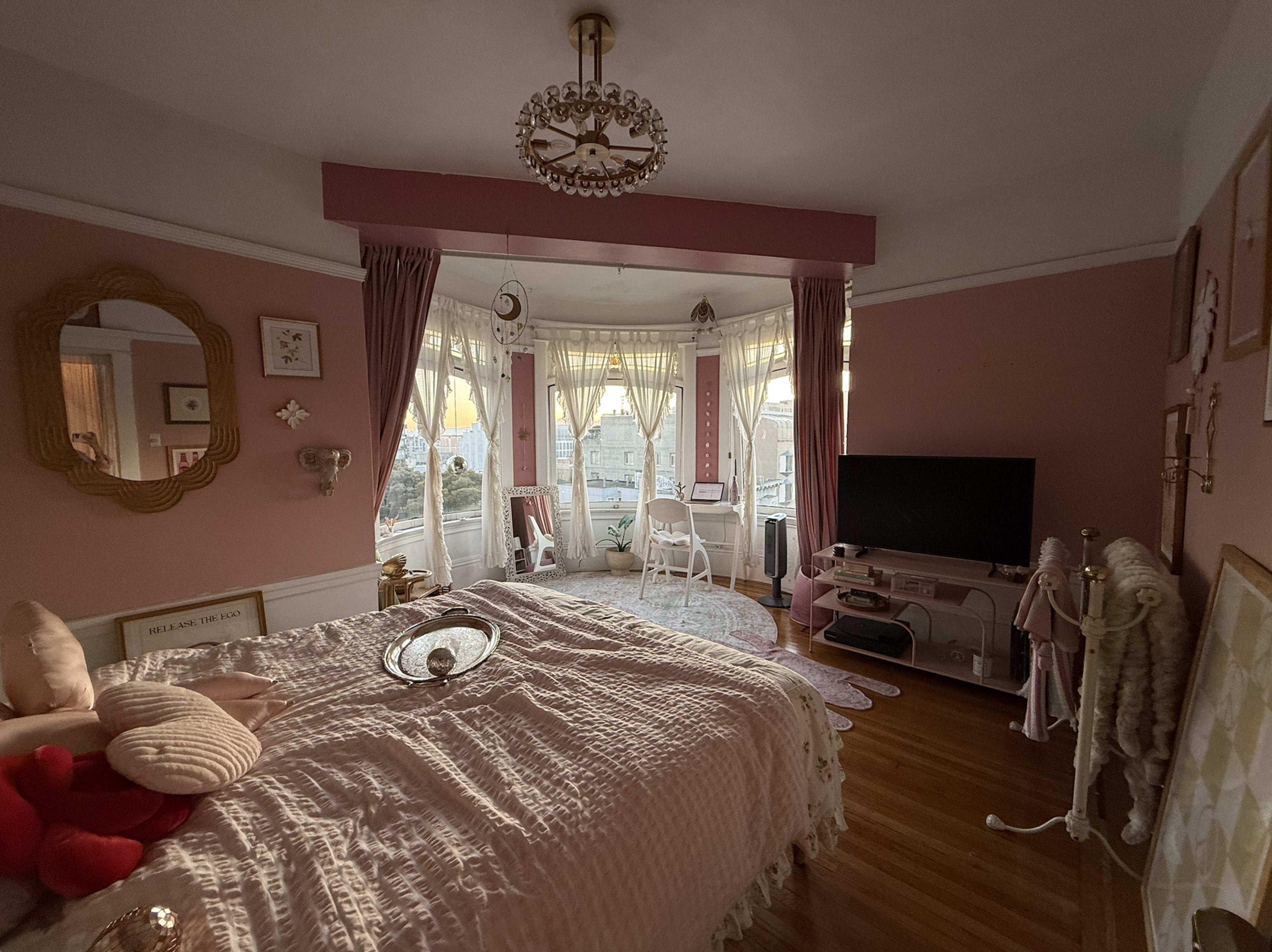 The image shows a cozy bedroom with a large bed, a TV stand, and multiple windows featuring sheer curtains that let in natural light, all adorned in soft pink hues.