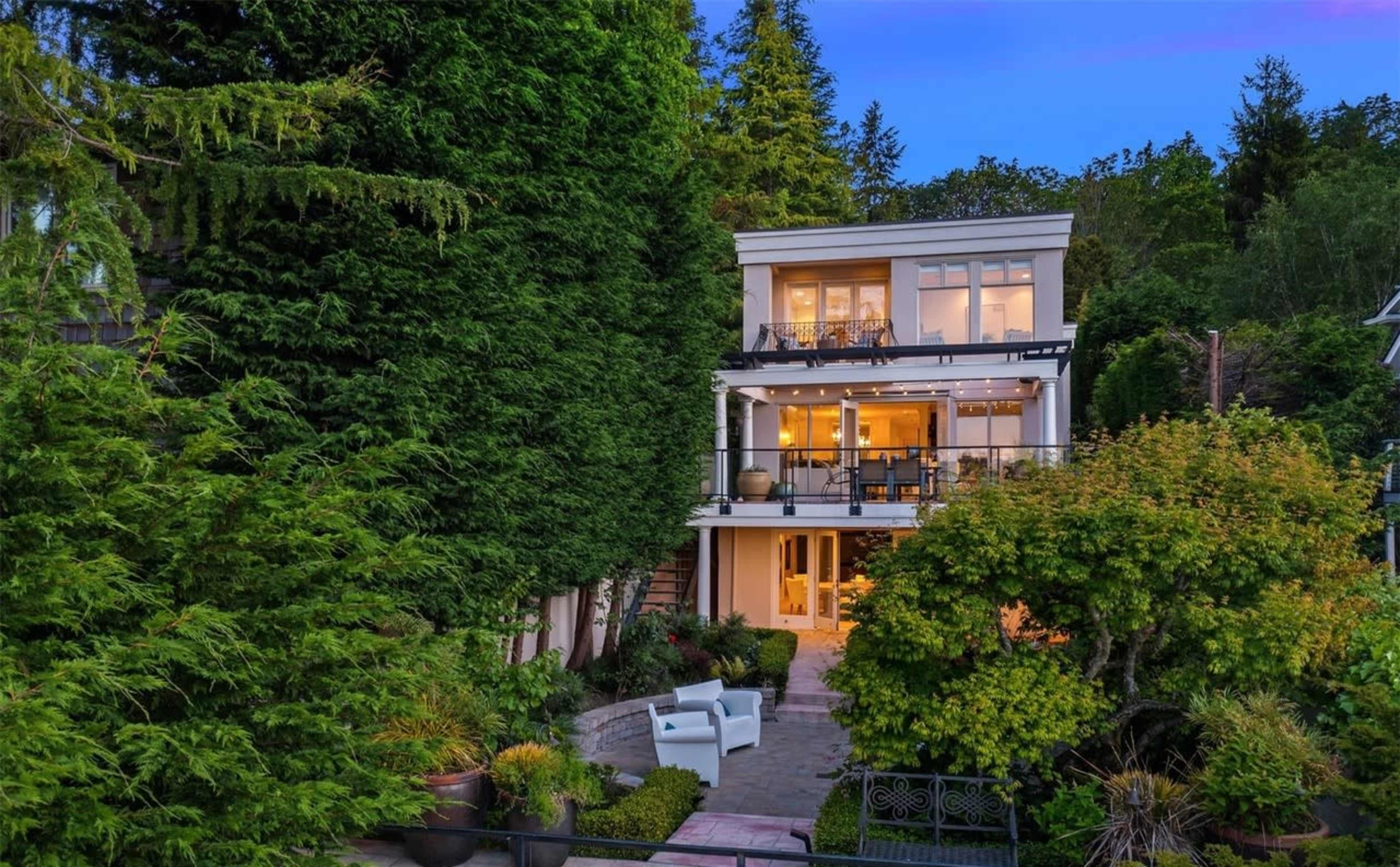 A multi-story house is surrounded by greenery, featuring large windows and a patio area in the front.