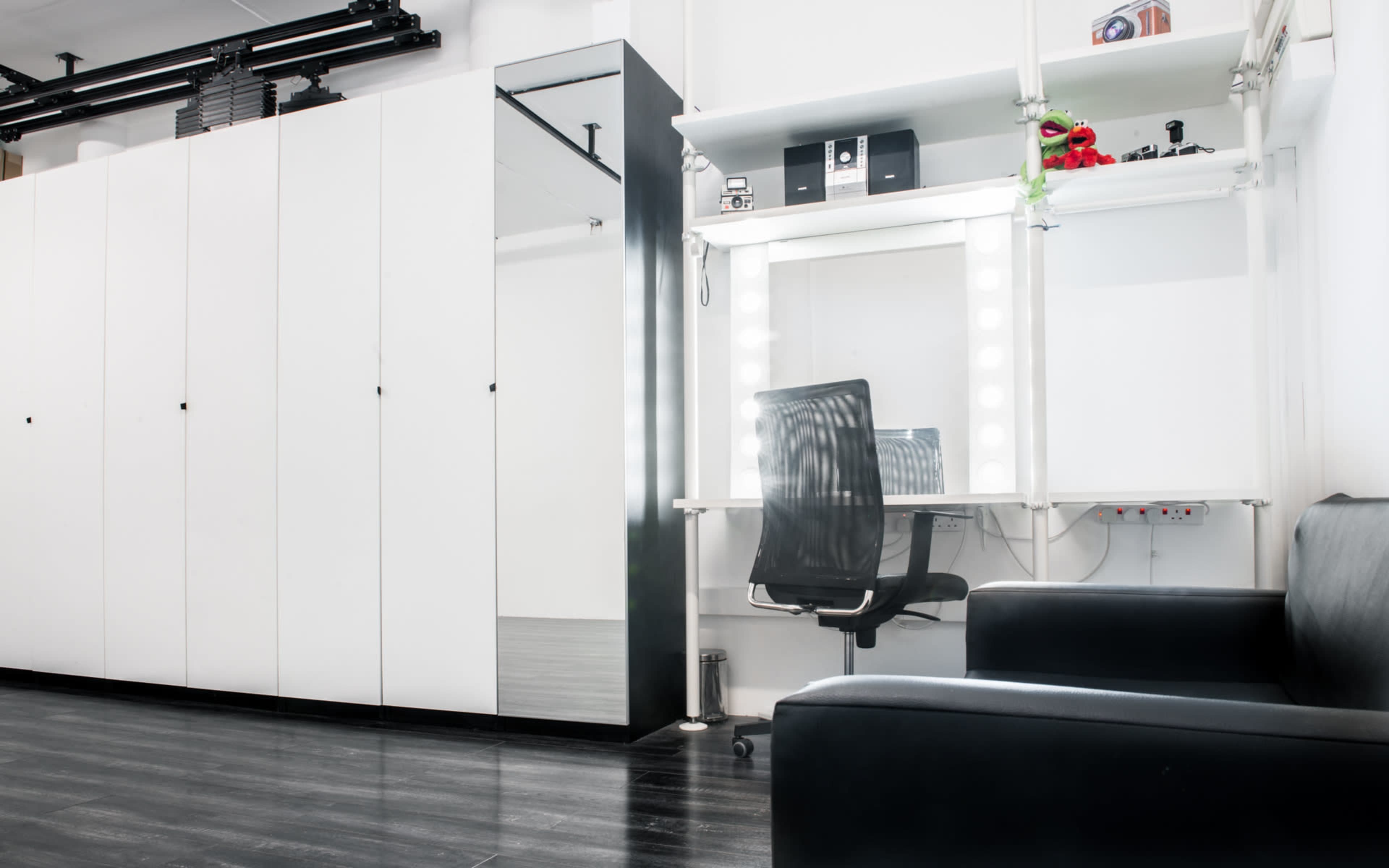 A minimalistic studio space featuring a black sofa, a mirrored wardrobe, and a well-lit vanity area with a chair and shelves.