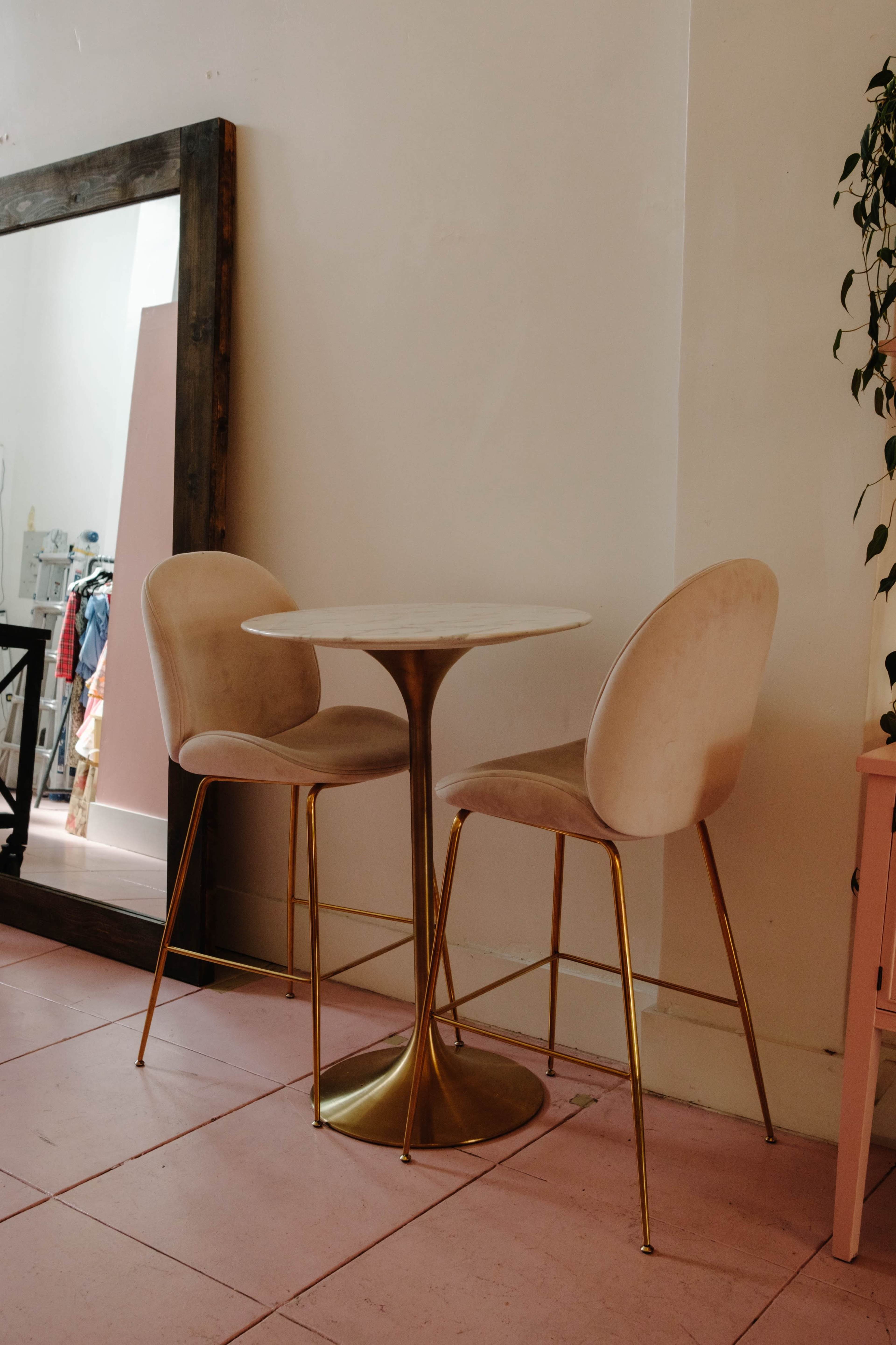 A small round table with a brass base and two upholstered chairs stands against a pale wall, next to a large mirror.