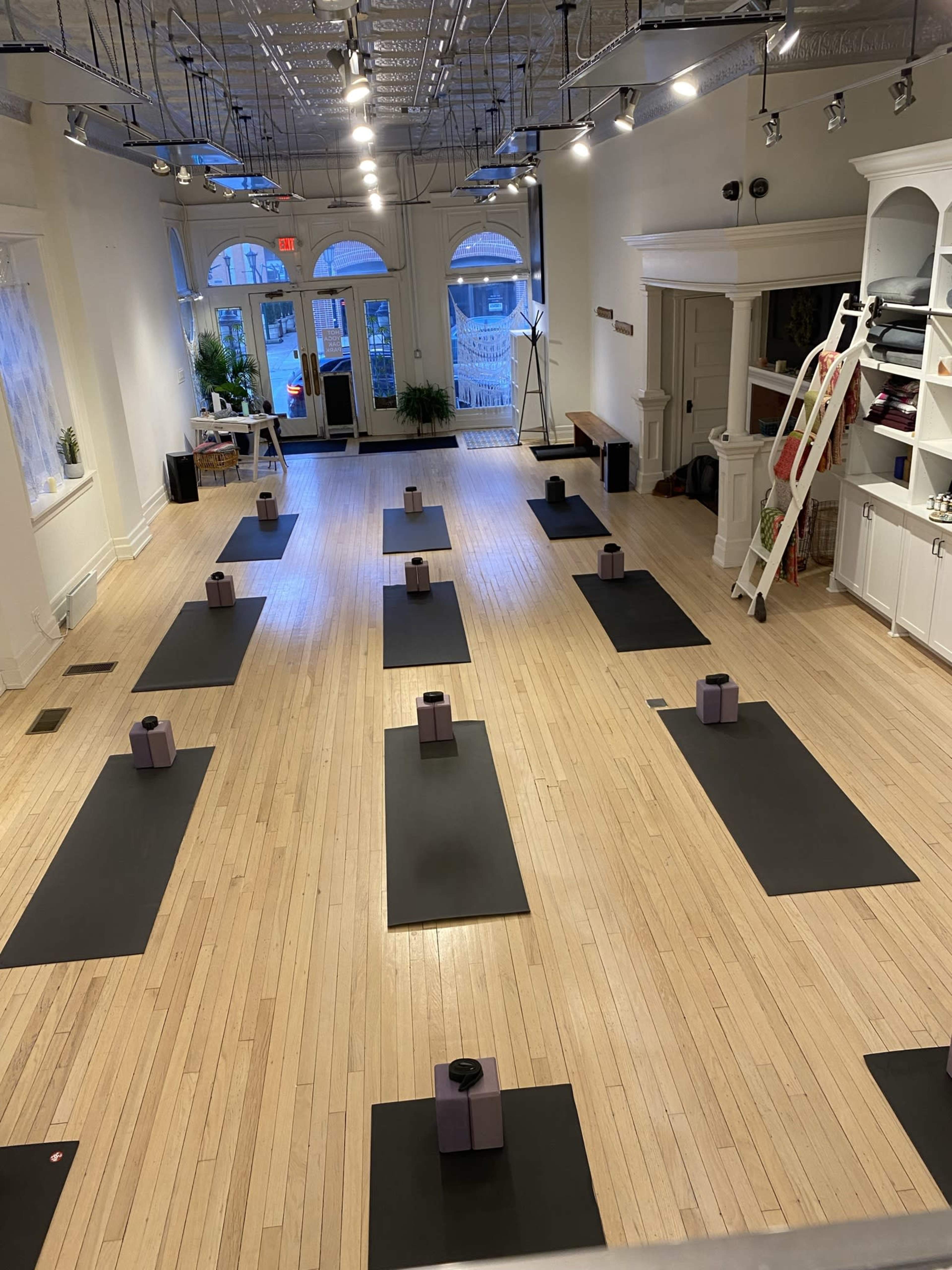 The image shows a spacious yoga studio with several black mats arranged in rows, each featuring a bolstered prop at the center, and large windows allowing natural light to enter.