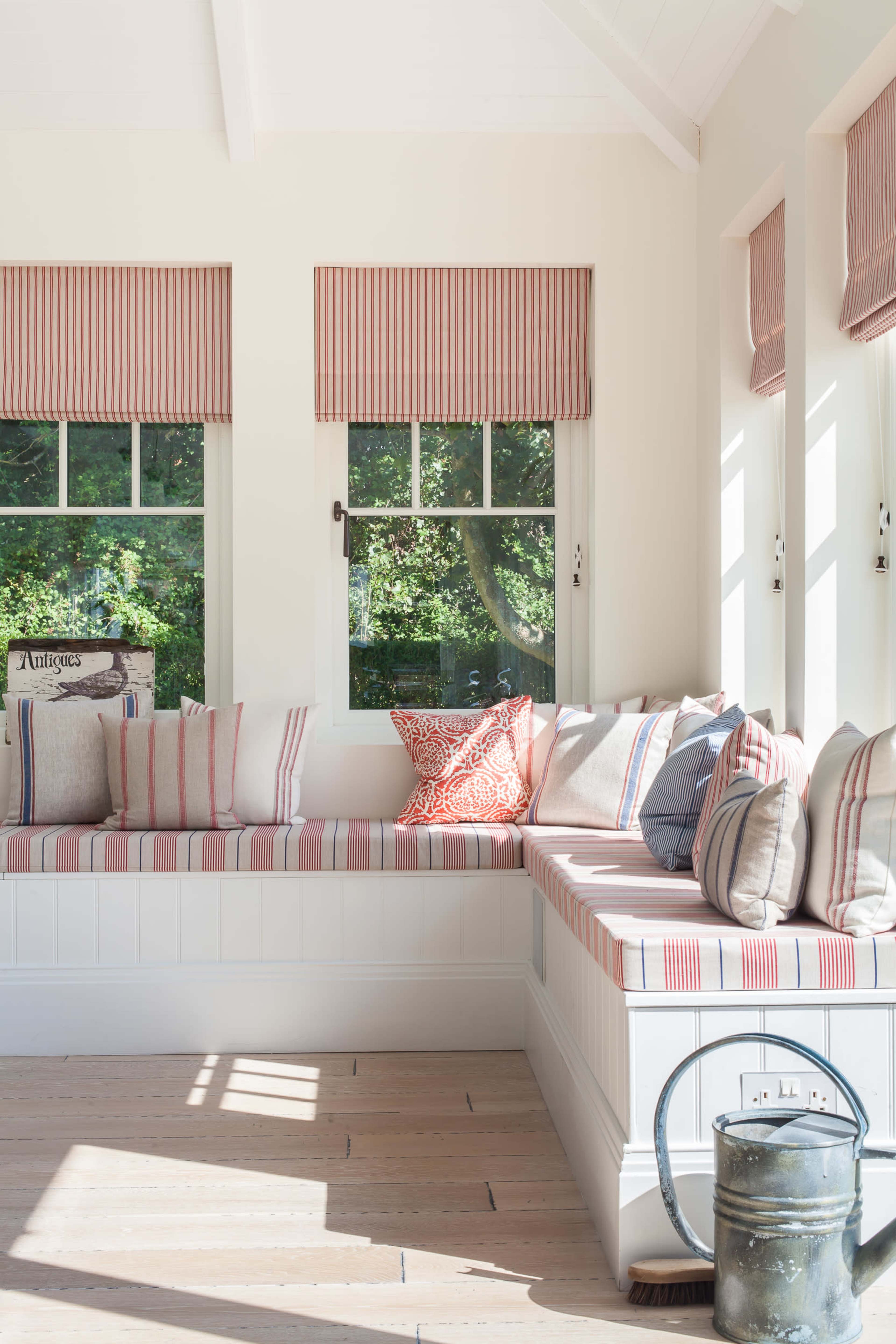 A bright sunroom with a built-in bench adorned with striped cushions and pillows, while sunlight streams in through the windows.