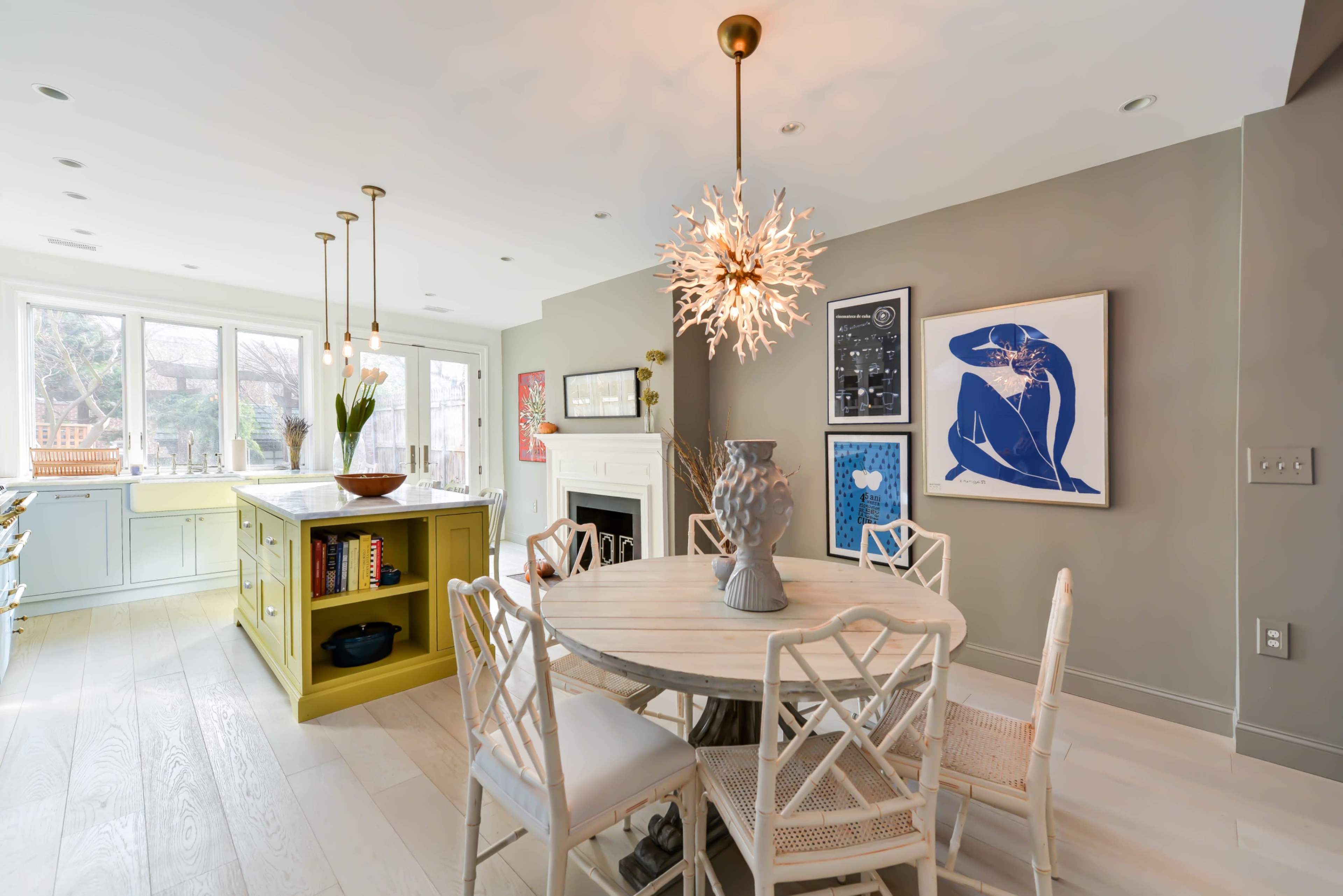 A modern dining area with a round table surrounded by six chairs, a vibrant yellow kitchen island, and a large window allowing natural light to illuminate the space.