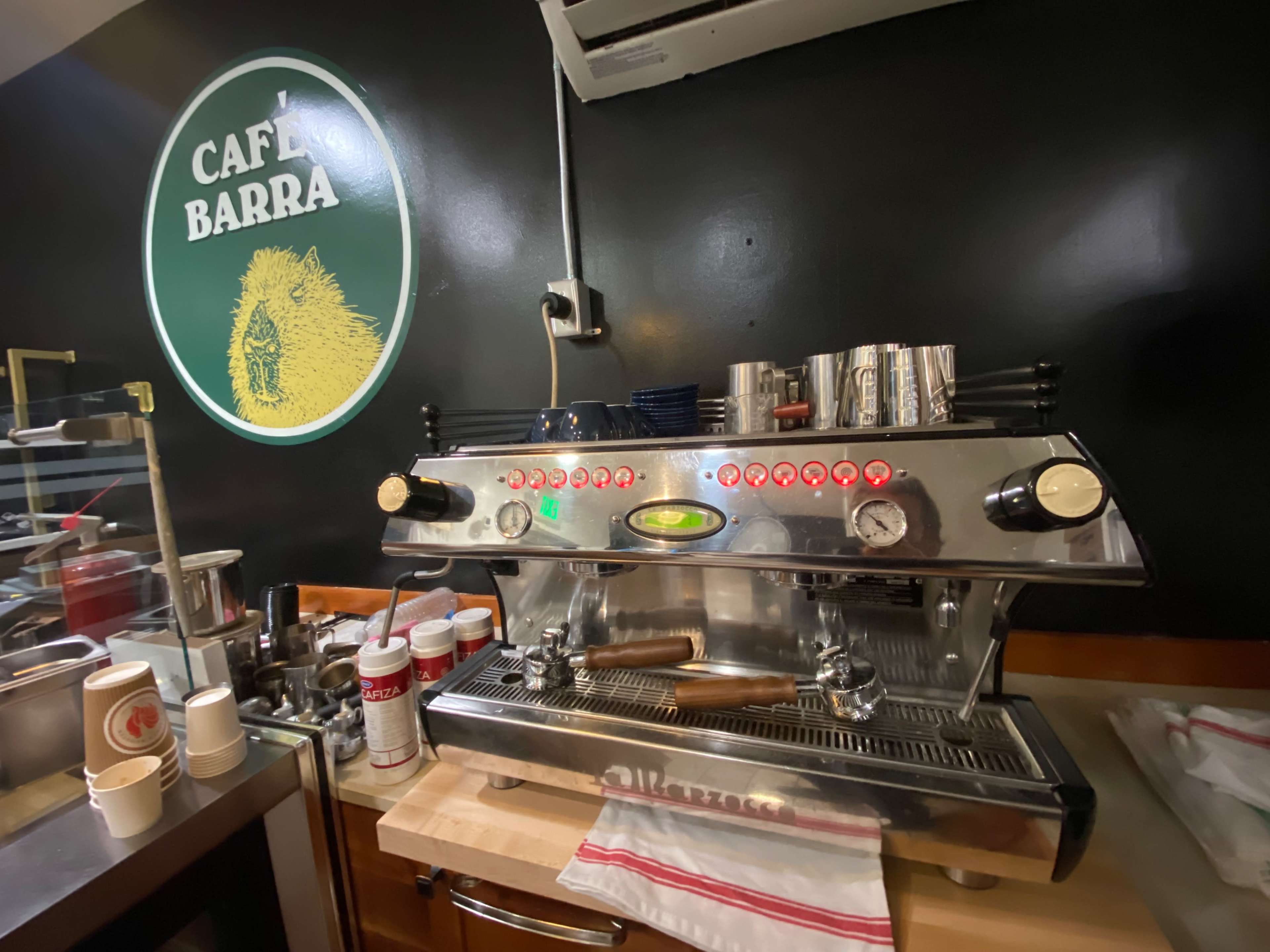 A shiny espresso machine is positioned on a countertop beneath a sign that reads "Café Barra."