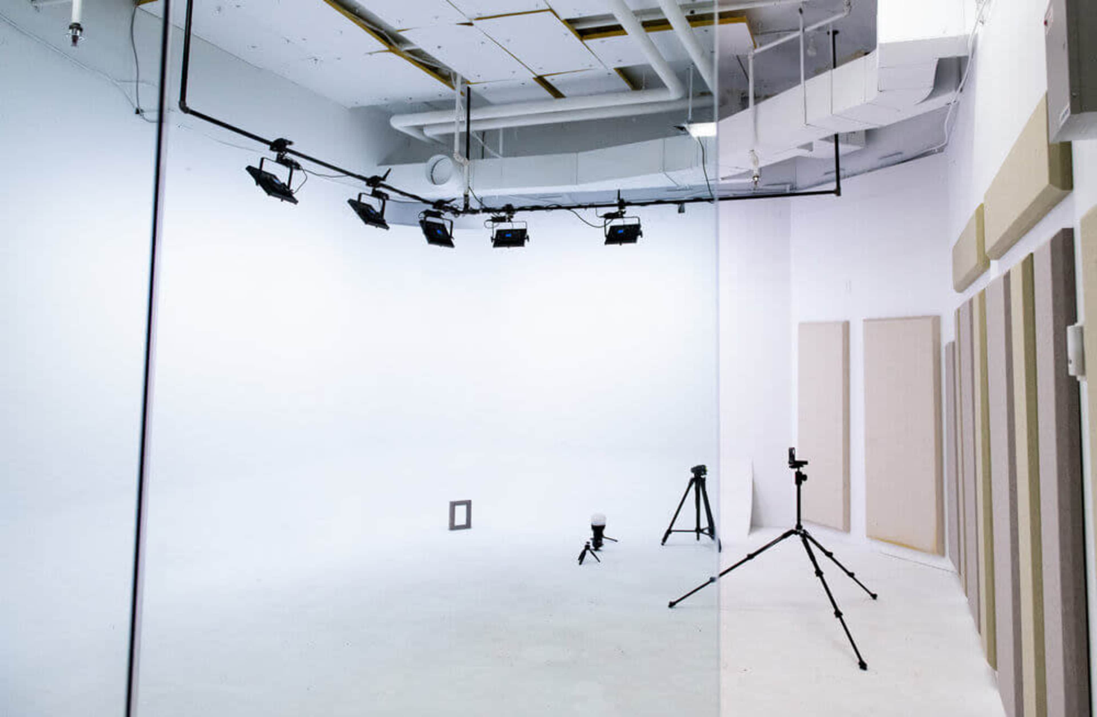 The image shows an empty studio with white walls, overhead lighting, and camera equipment set up on tripods.