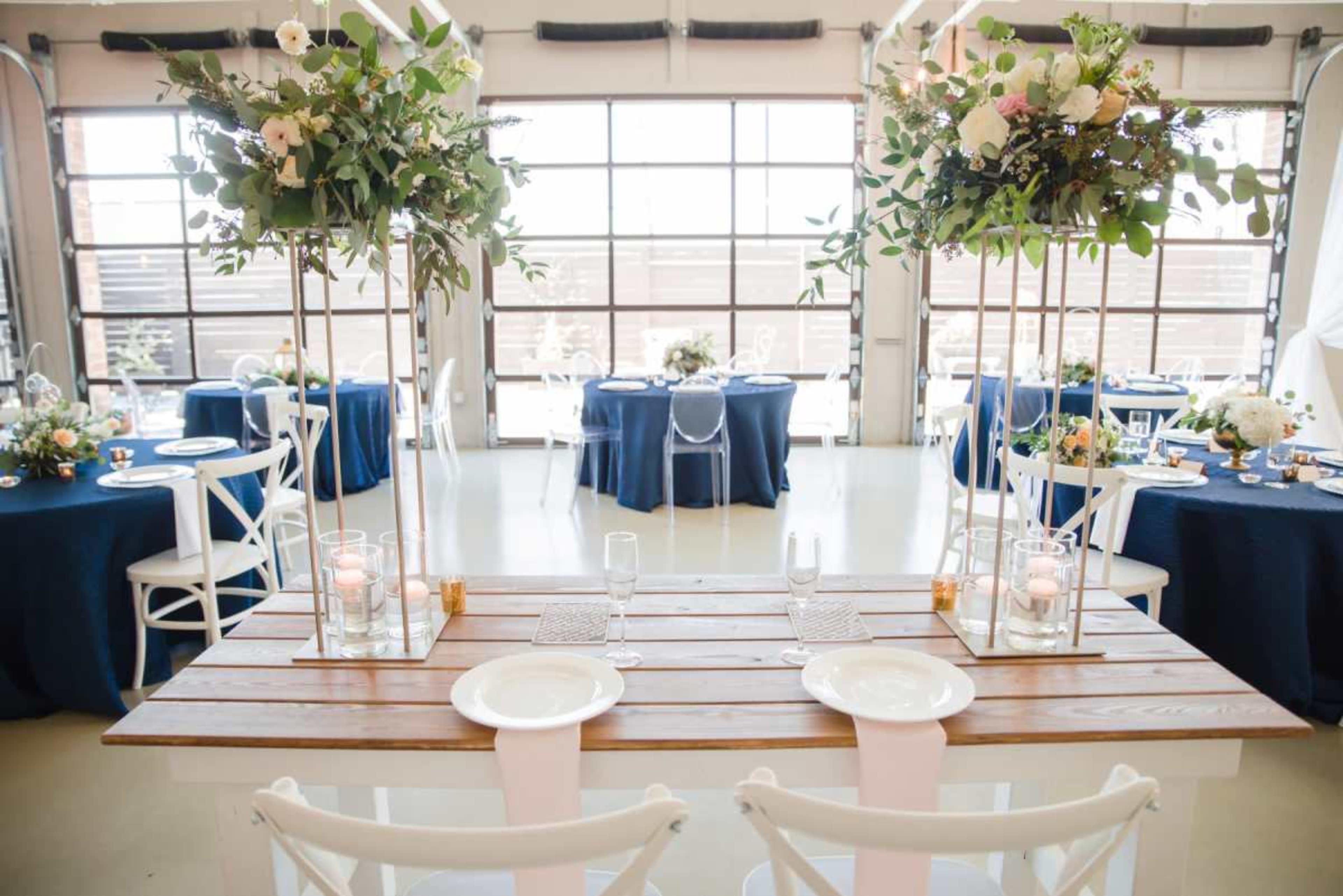 A decorated banquet space features wooden tables with floral arrangements, navy blue tablecloths, and transparent chairs set for an event.