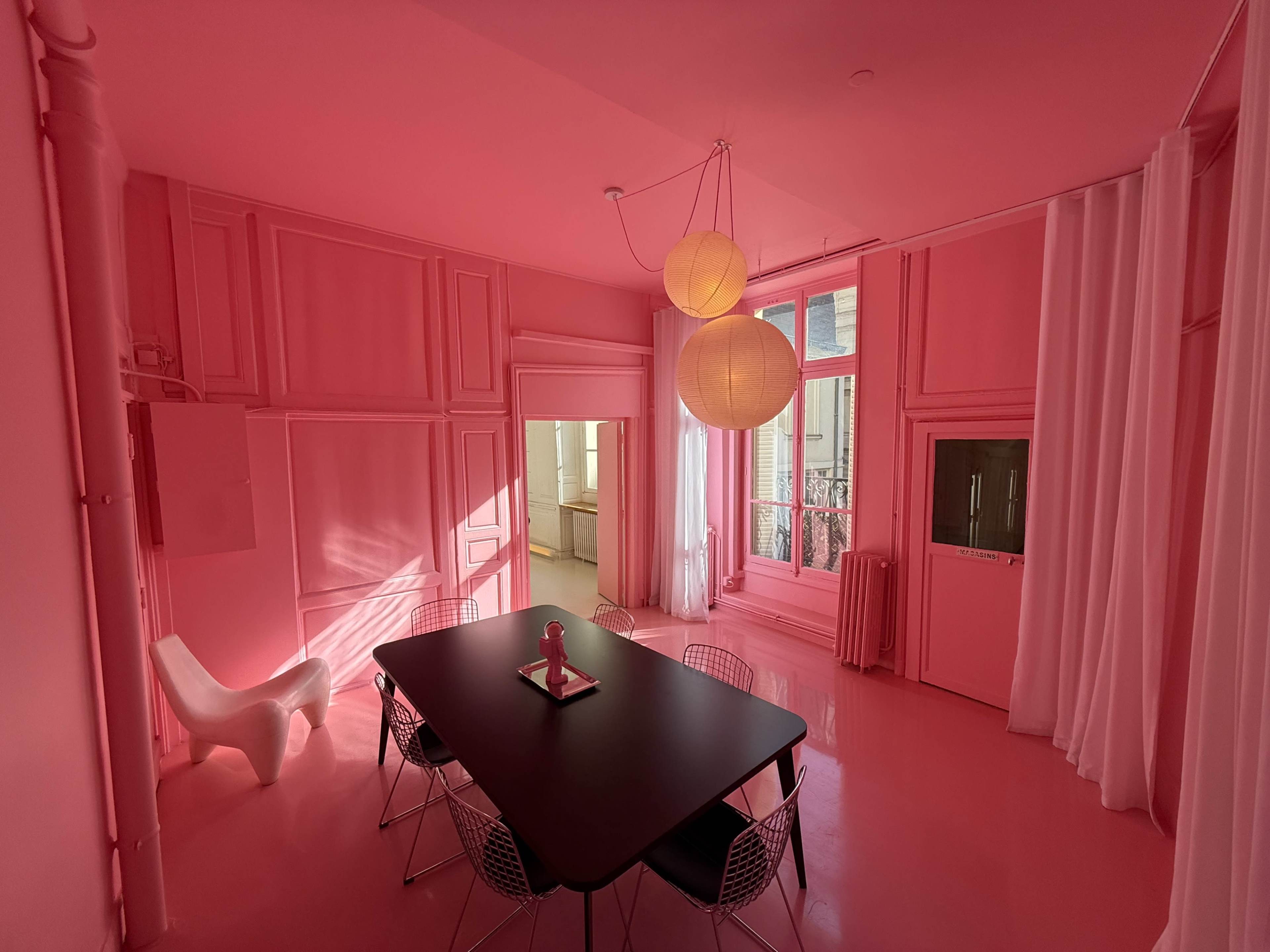 The image shows a fully pink room featuring a black dining table surrounded by wire chairs, with large windows allowing natural light to enter.