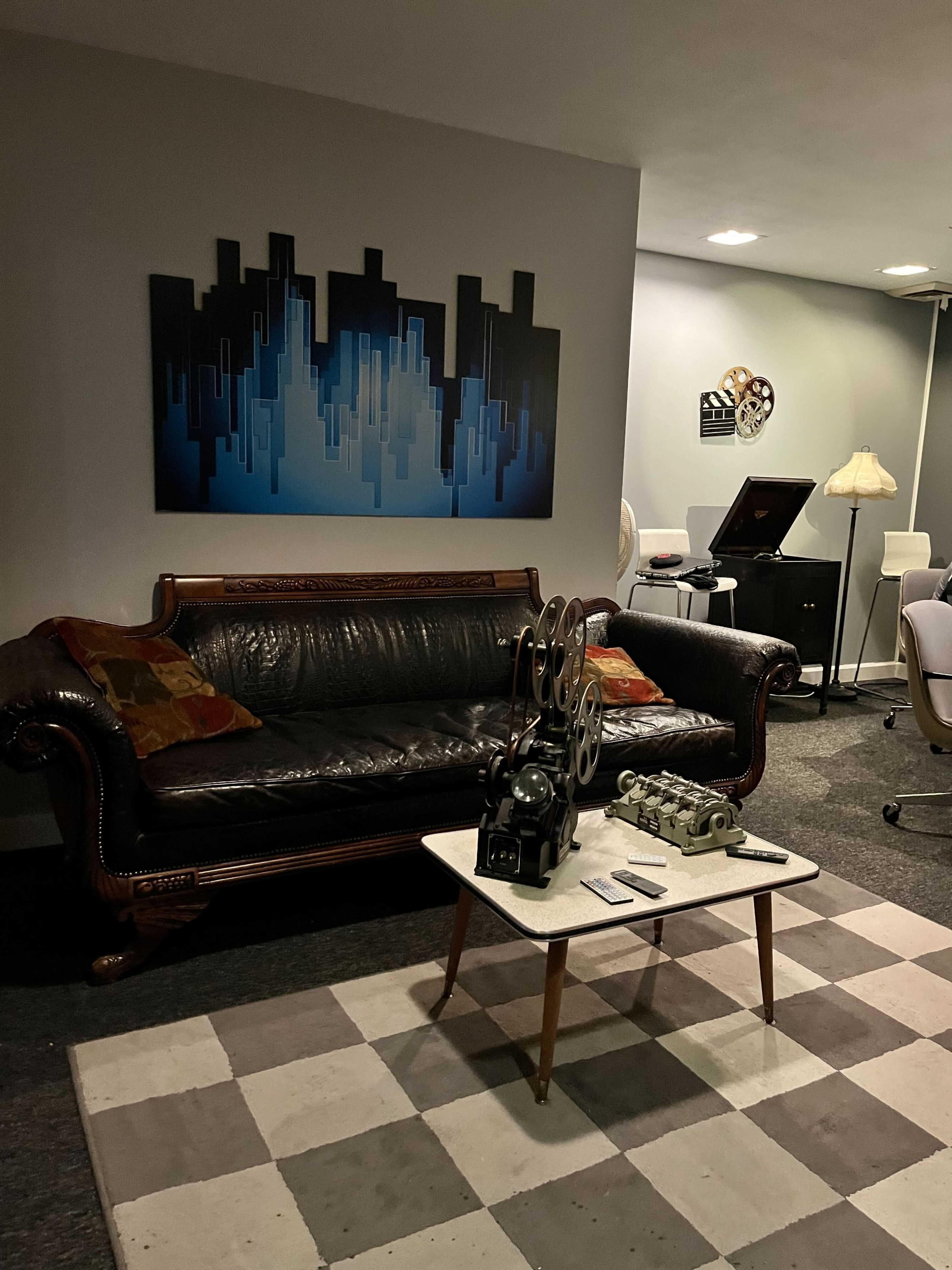 The image shows a cozy basement living area with a black leather couch, a patterned rug, a coffee table with film projectors, and a wall art piece depicting a city skyline.