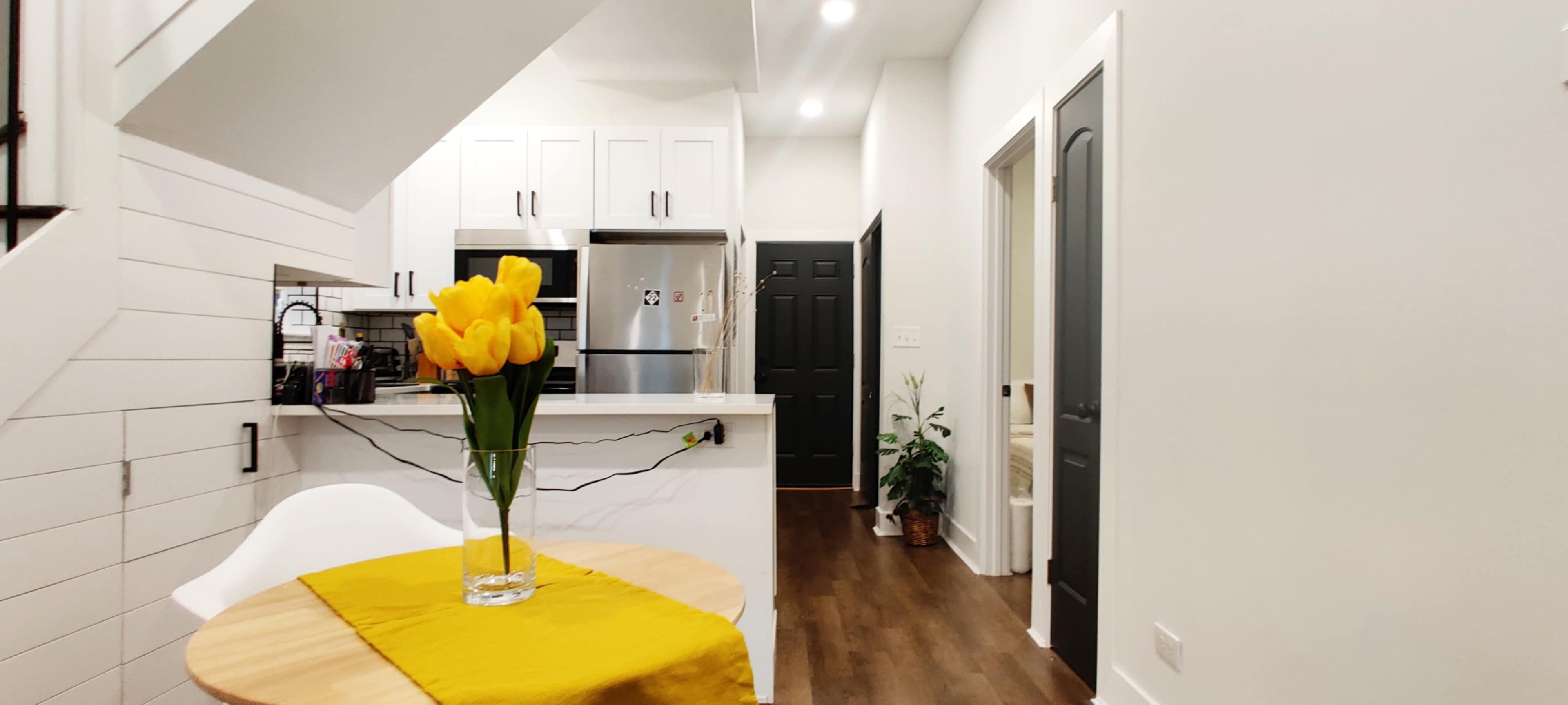 A modern kitchen and hallway are illuminated, featuring a vase of yellow tulips on a wooden table and black doors leading to the rest of the home.