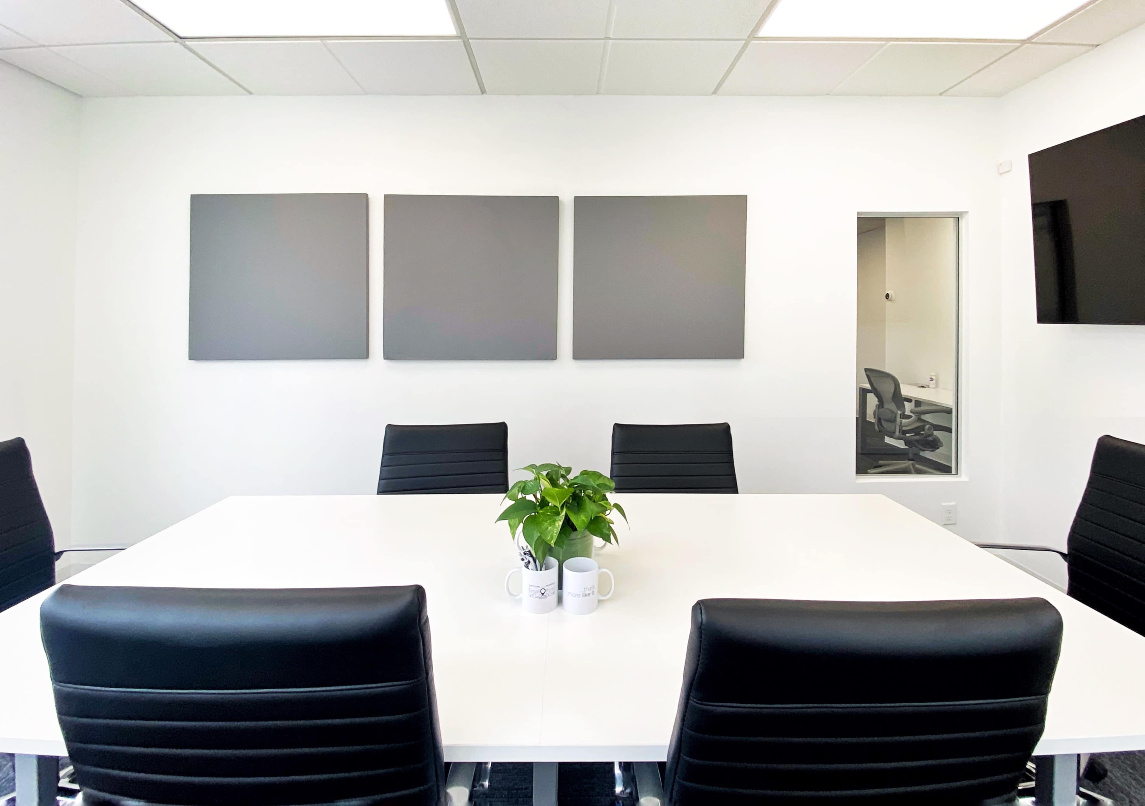 A modern conference room features a large white table surrounded by black chairs, with three blank gray canvases on the wall and a small potted plant in the center.