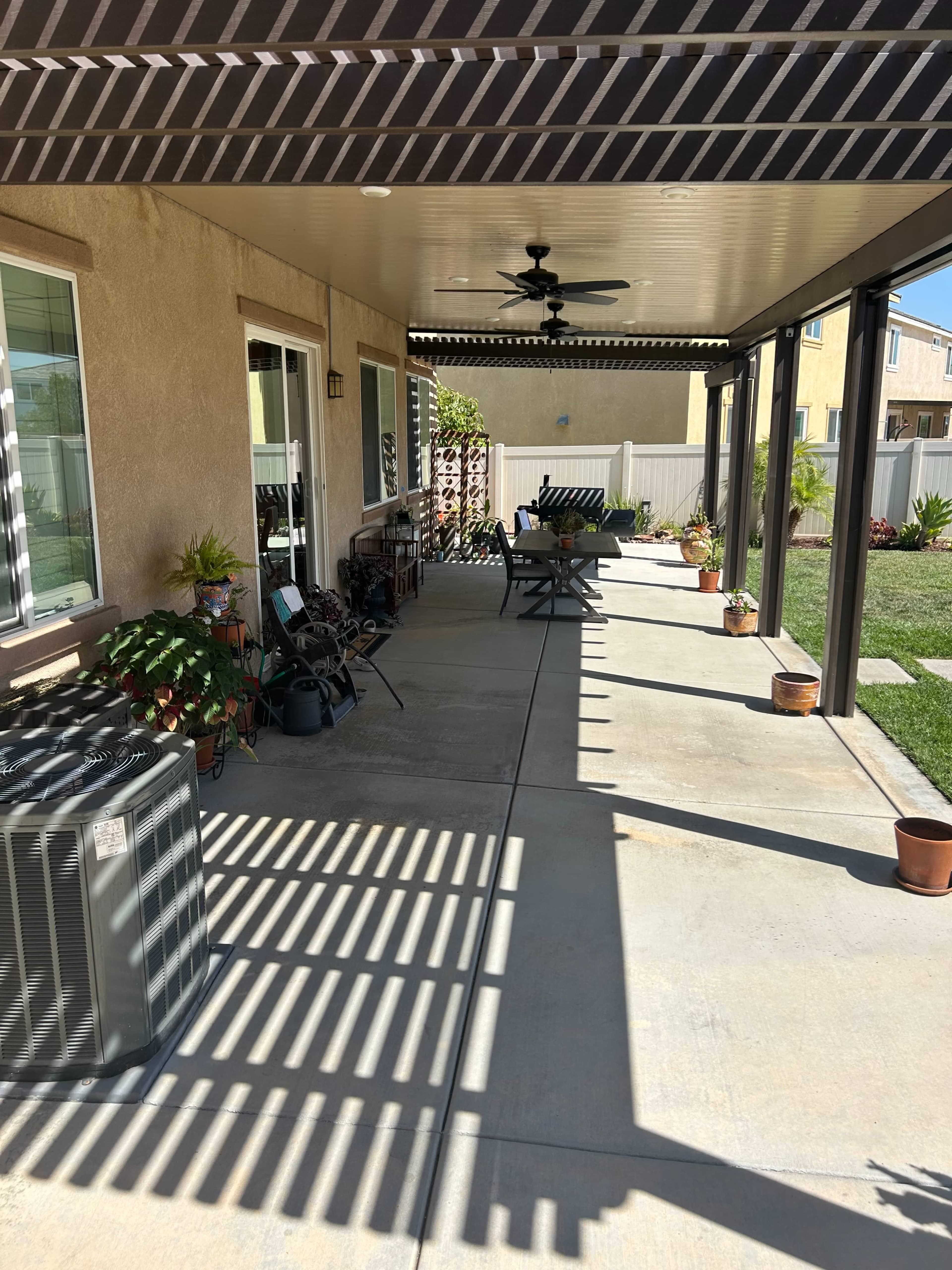 The image shows a covered patio area featuring a concrete floor, potted plants, outdoor furniture, and an air conditioning unit along the side.