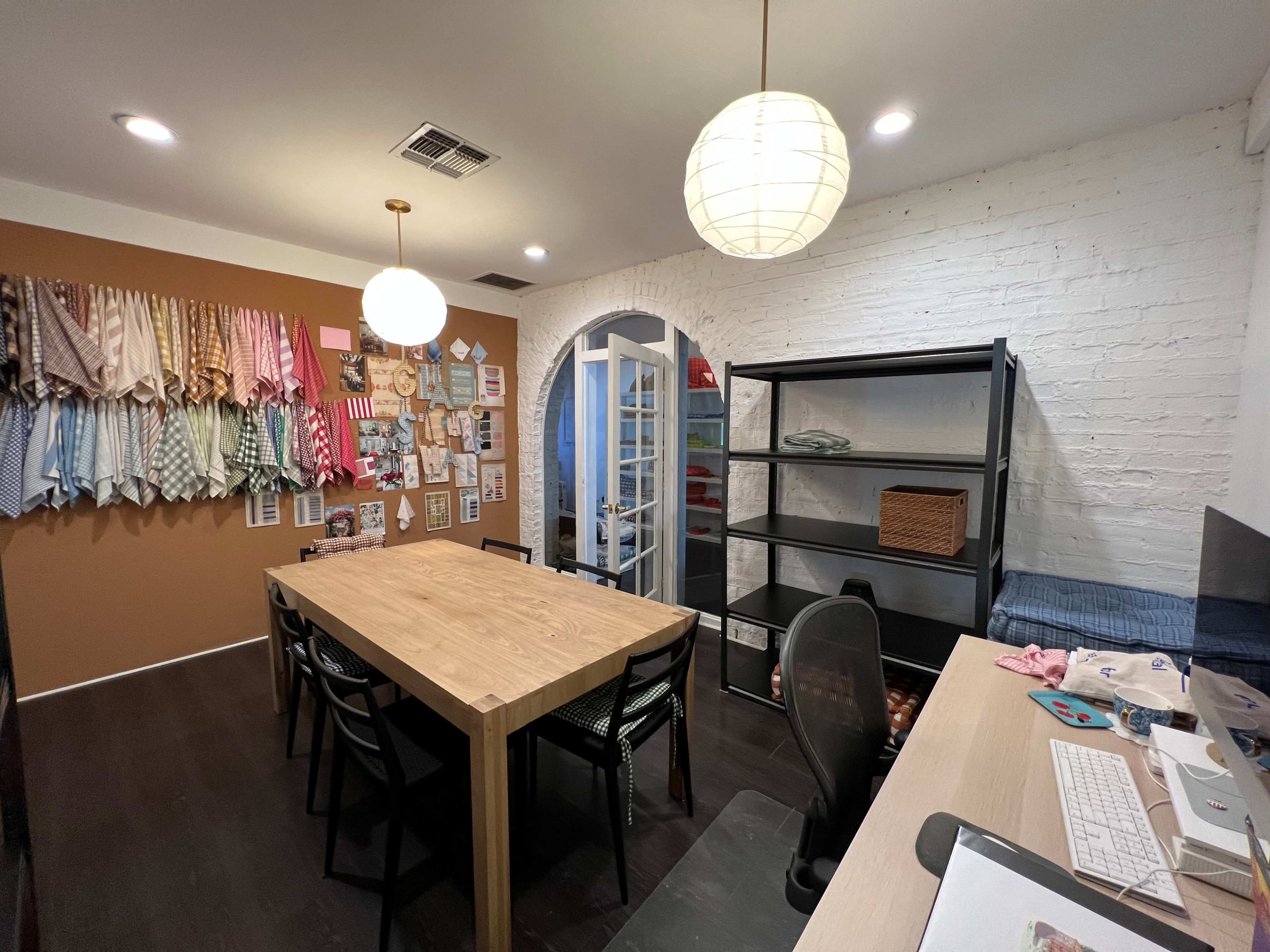 A well-organized workspace features a large wooden table, a display of fabric samples on the wall, and a shelving unit alongside a glass door leading to another area.
