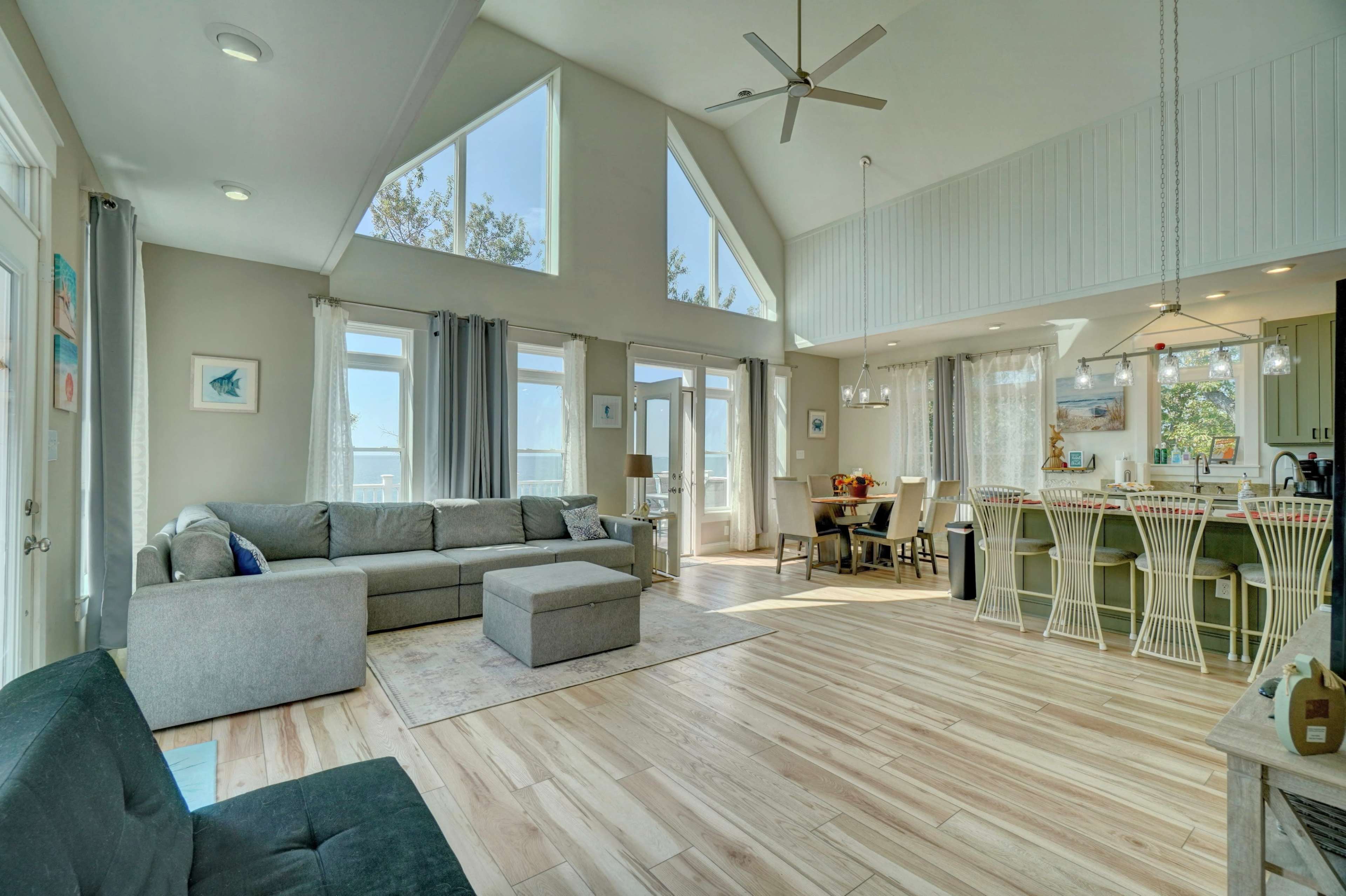 The image shows a spacious, modern living area with large windows, wooden flooring, and a mix of seating arrangements including a sofa and dining area.