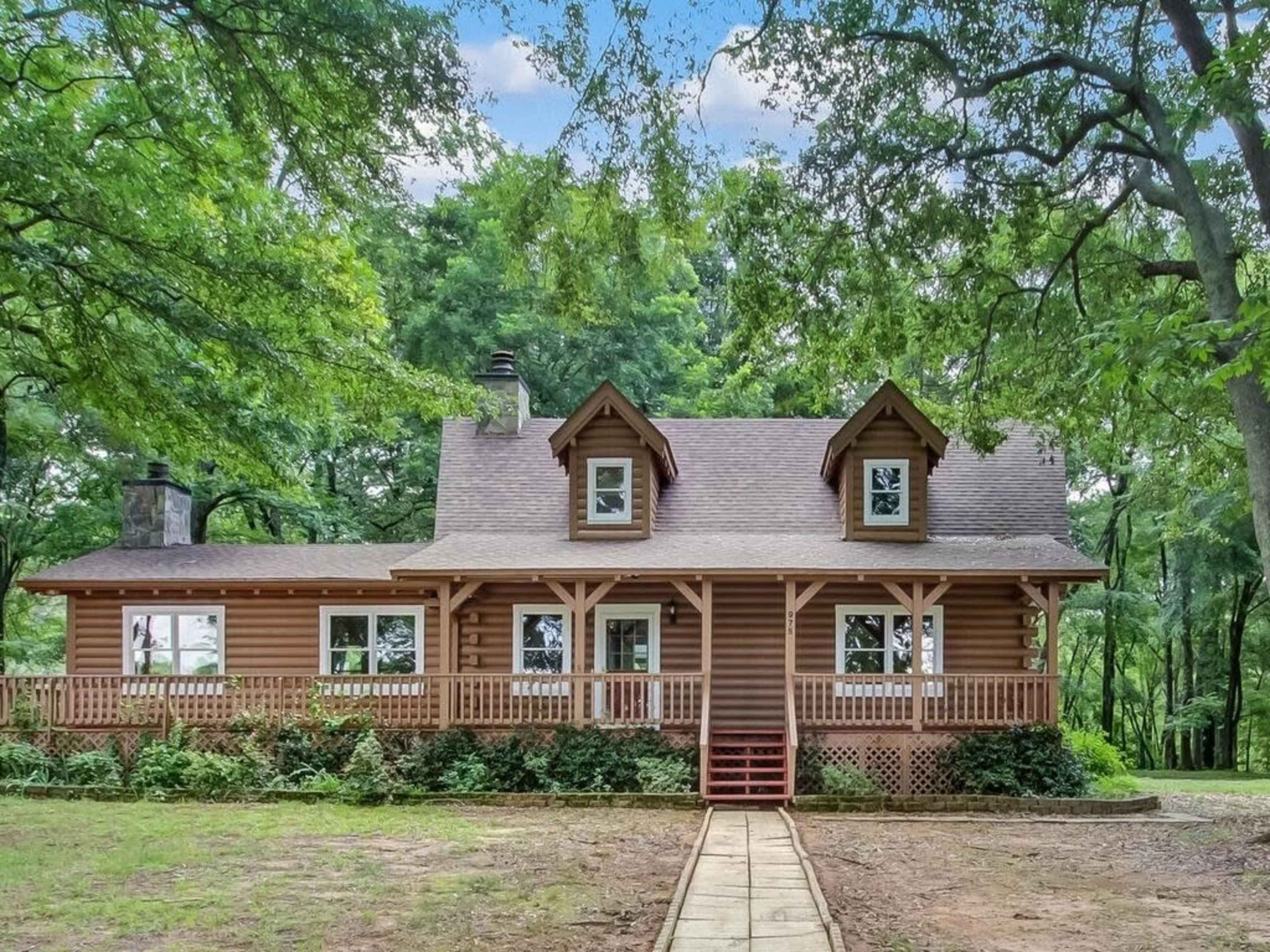 A log cabin-style house with a porch is surrounded by lush greenery and trees.