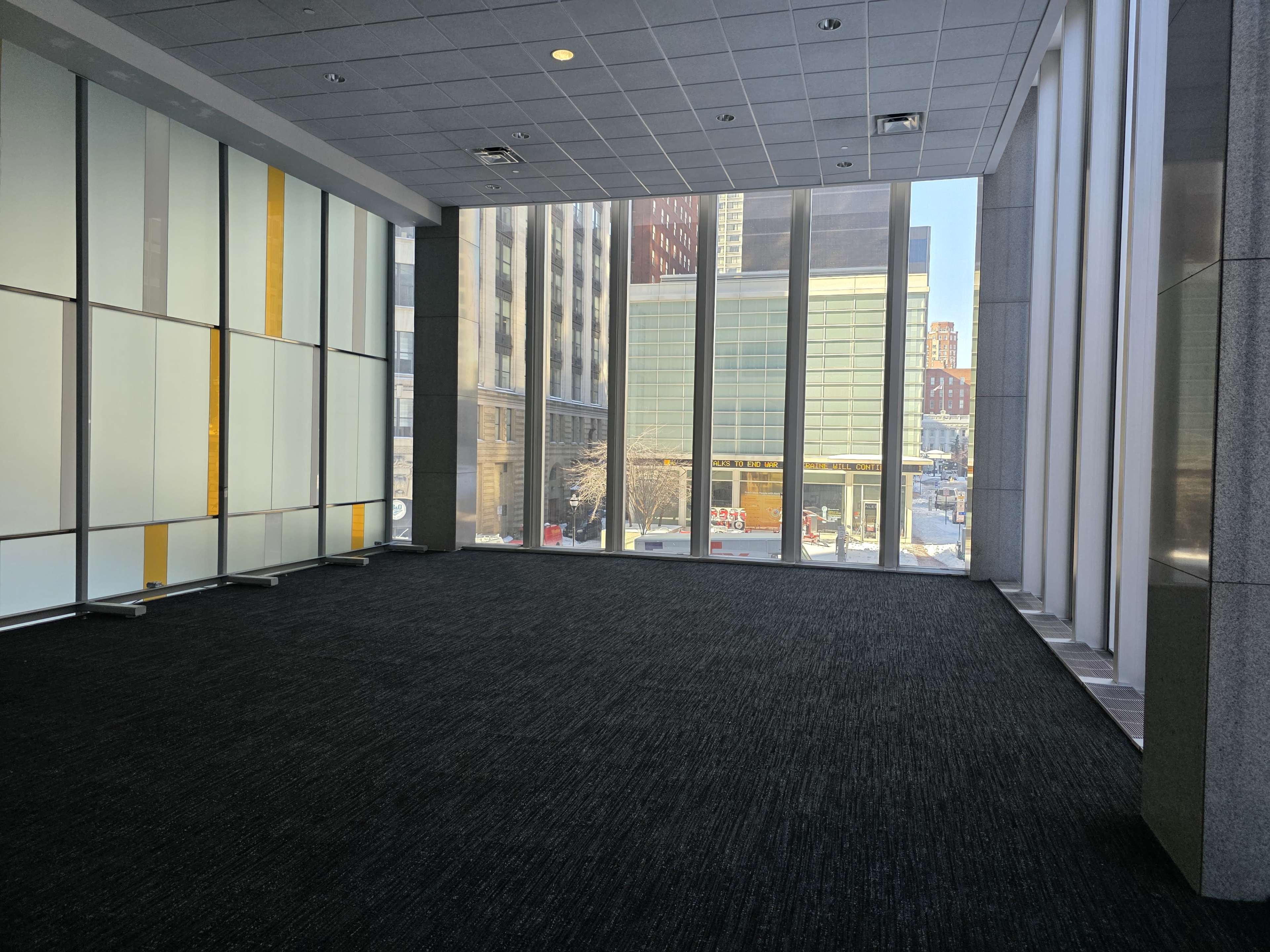 The image shows an empty room with large windows overlooking a cityscape, featuring a carpeted floor and walls with glass panels.