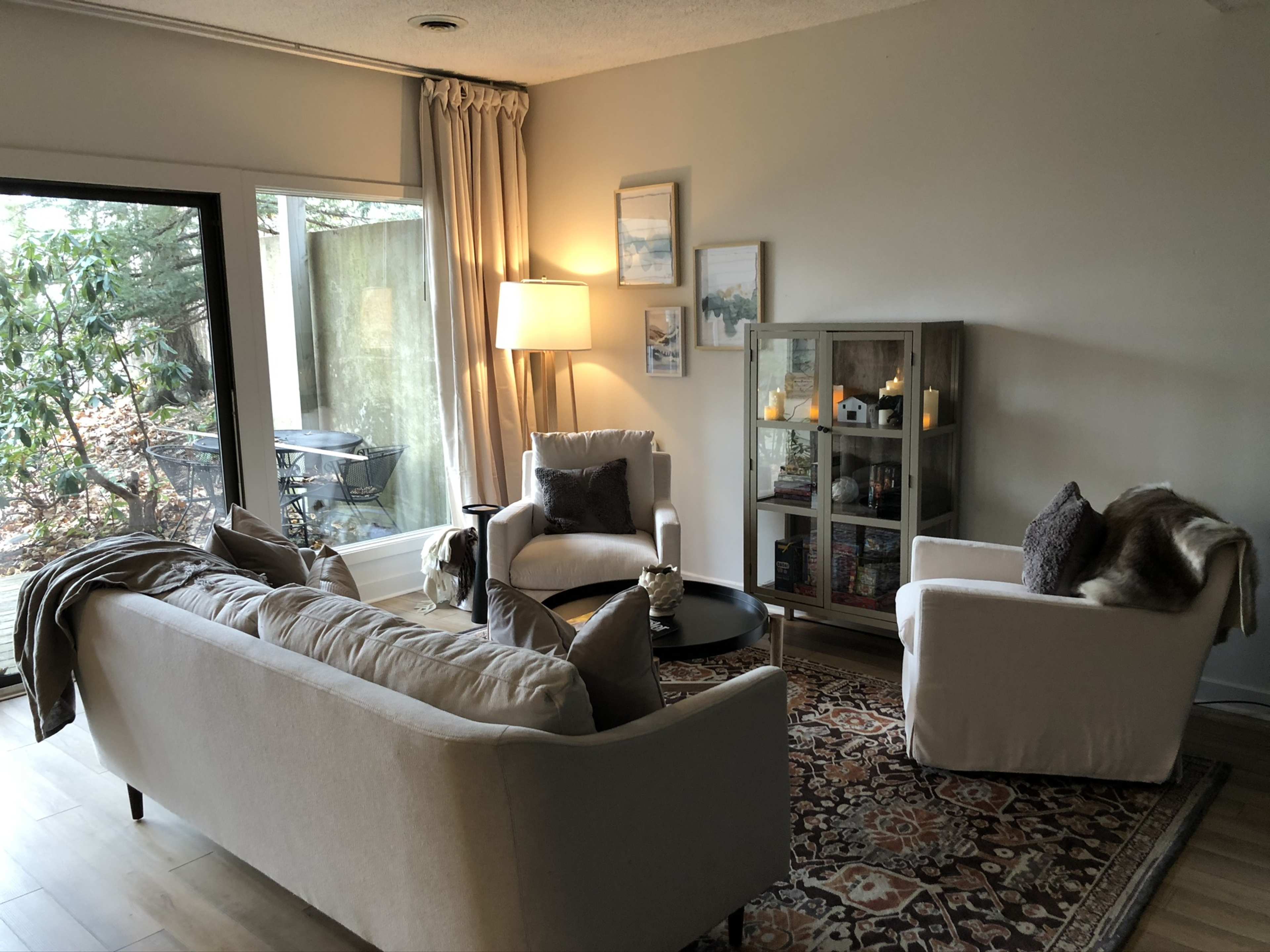 A cozy living room features a cream-colored sofa, two armchairs, a round black coffee table, and a glass display cabinet, with a large window allowing natural light to enter.