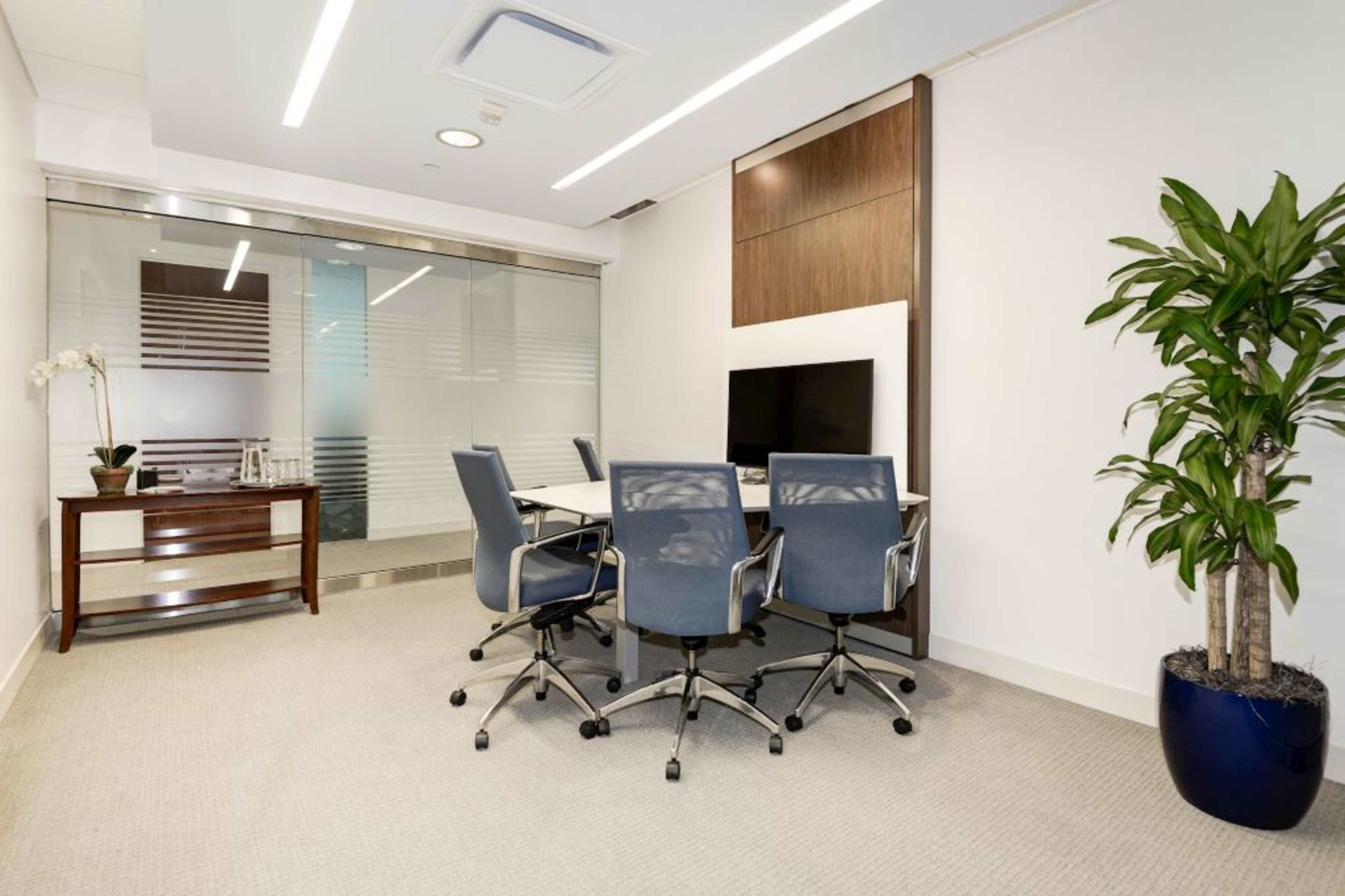 A modern conference room features a glass wall, a rectangular table with four blue chairs, and a kitchenette in the background.