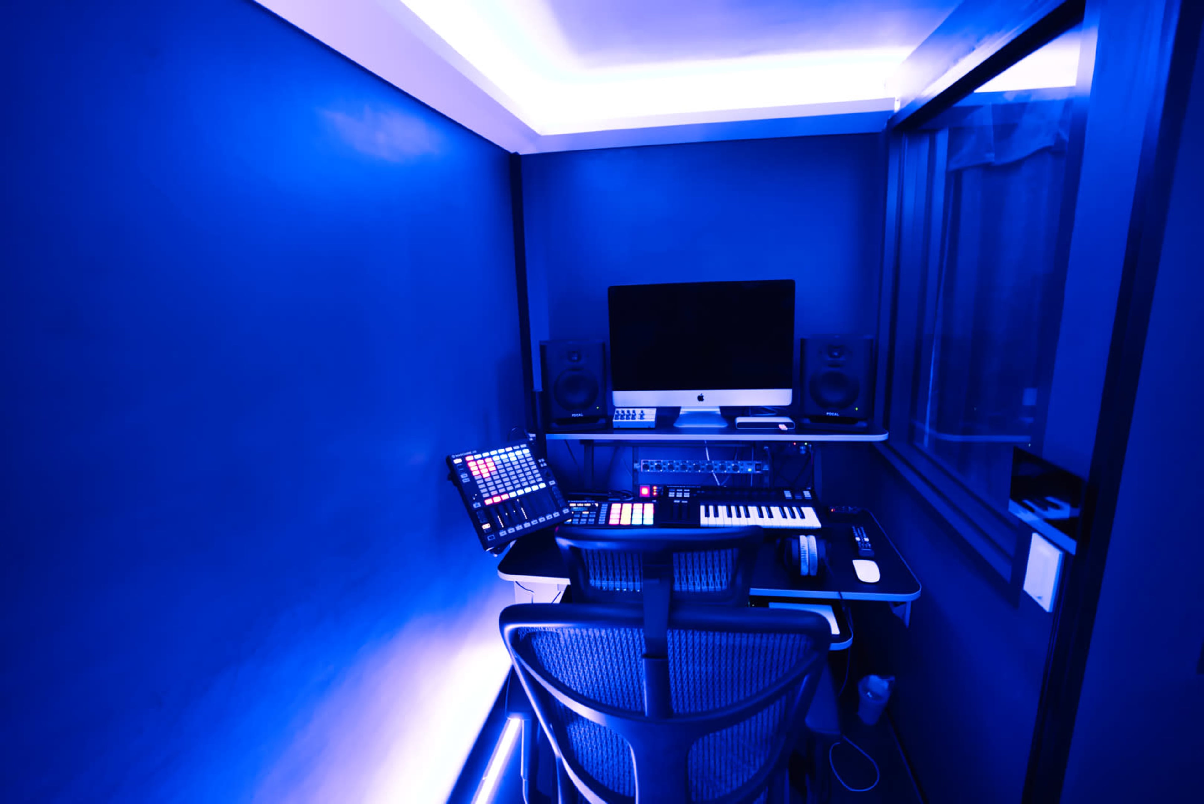A music production studio features a computer, MIDI keyboard, and sound equipment set against blue walls.
