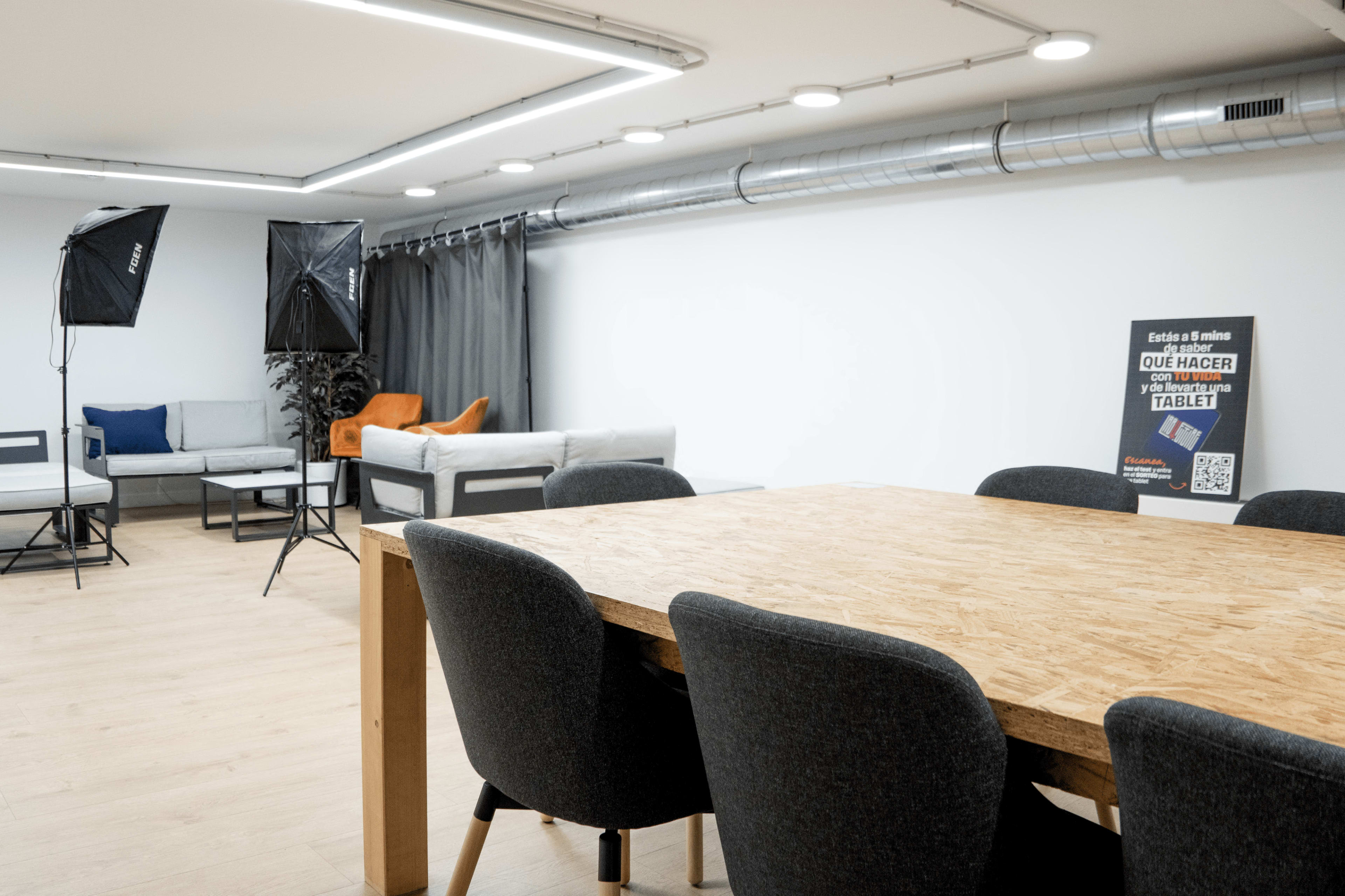 The image shows a modern workspace featuring a large wooden table surrounded by upholstered chairs, with sofas in the background and studio lighting equipment set up.
