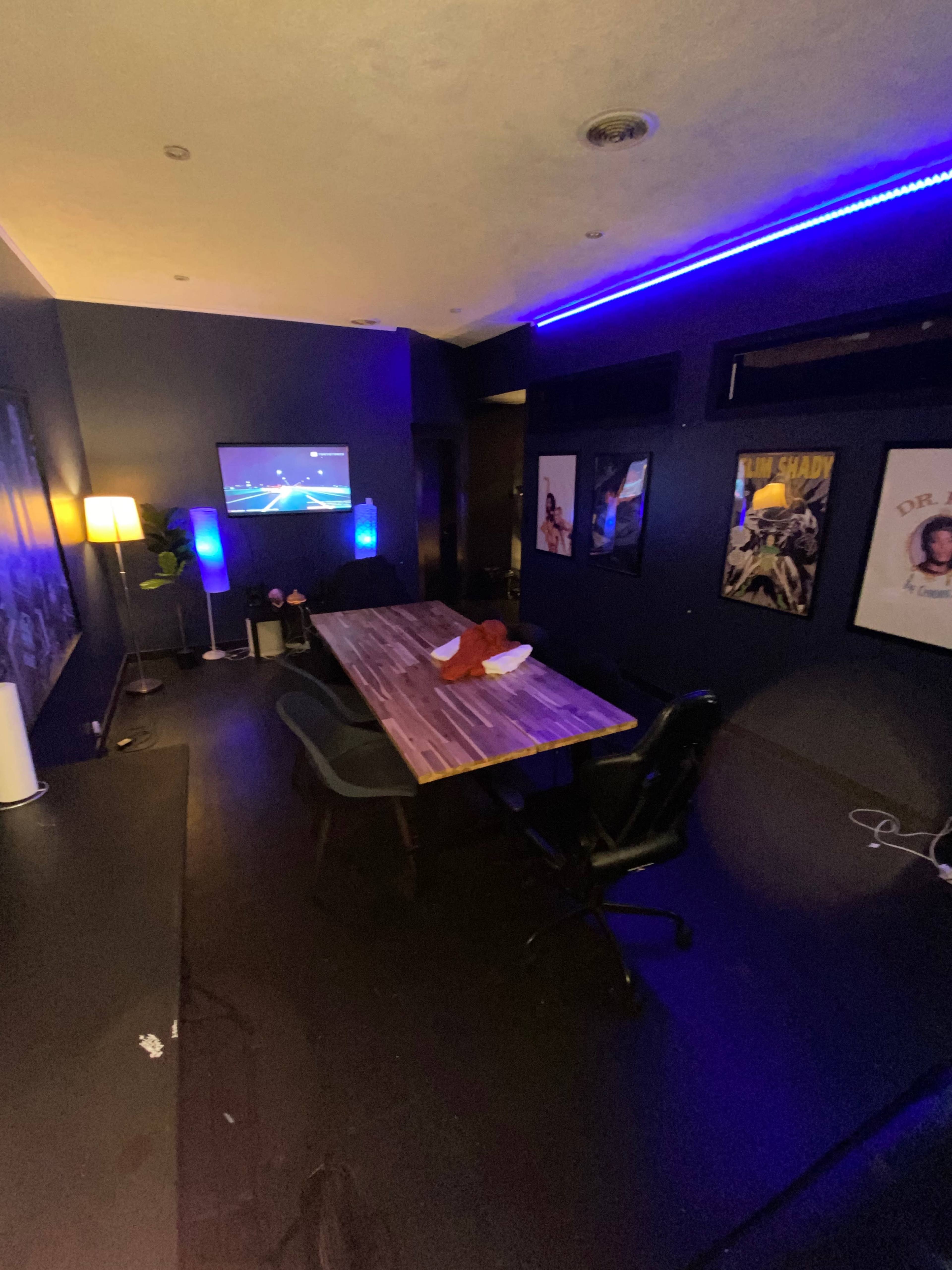A dimly lit room with a large table, chairs, a wall-mounted TV, and framed posters on the walls.