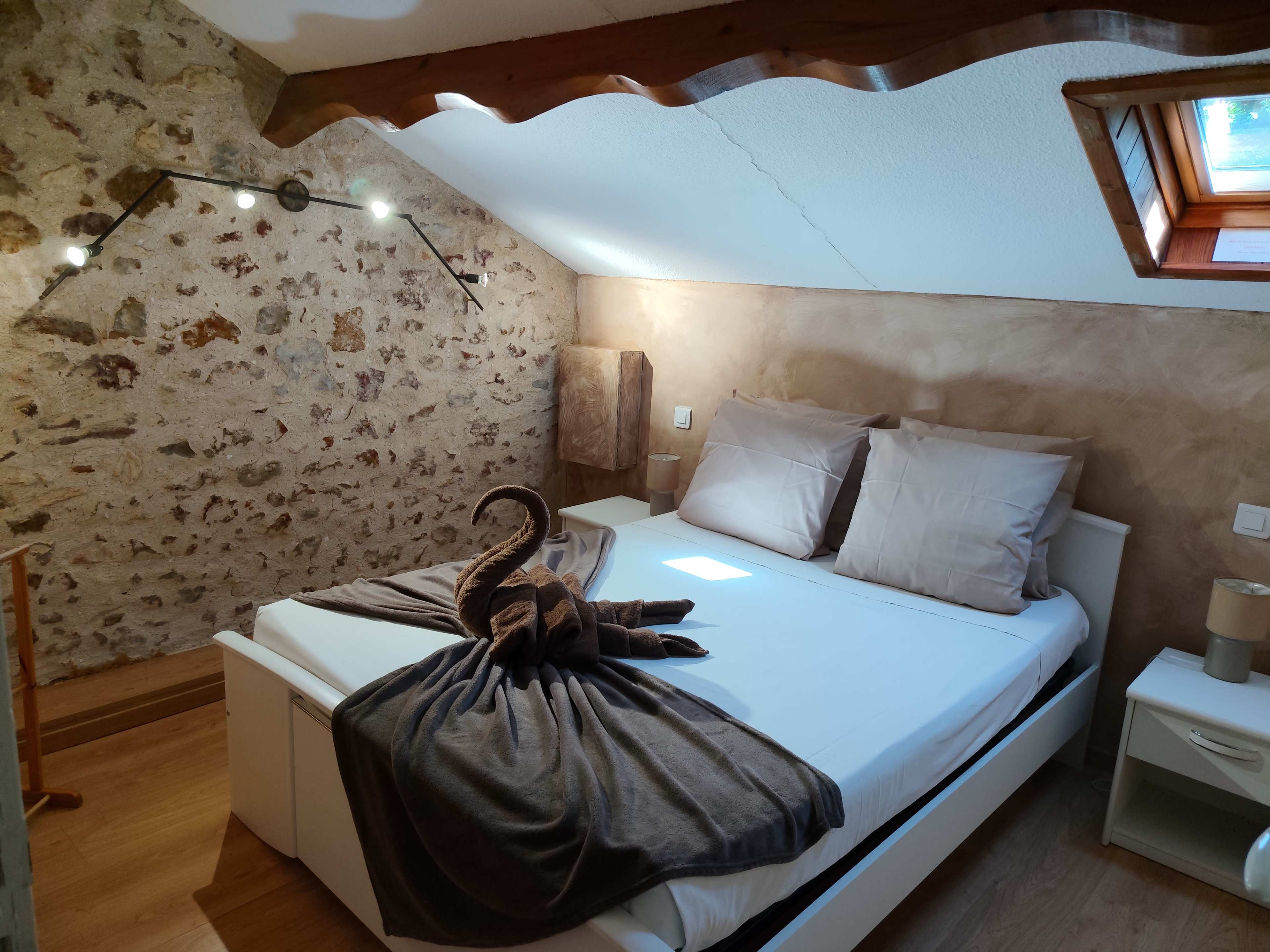 The image shows a cozy bedroom with a stone wall, featuring a bed adorned with beige pillows and a decorative towel shaped like a swan.