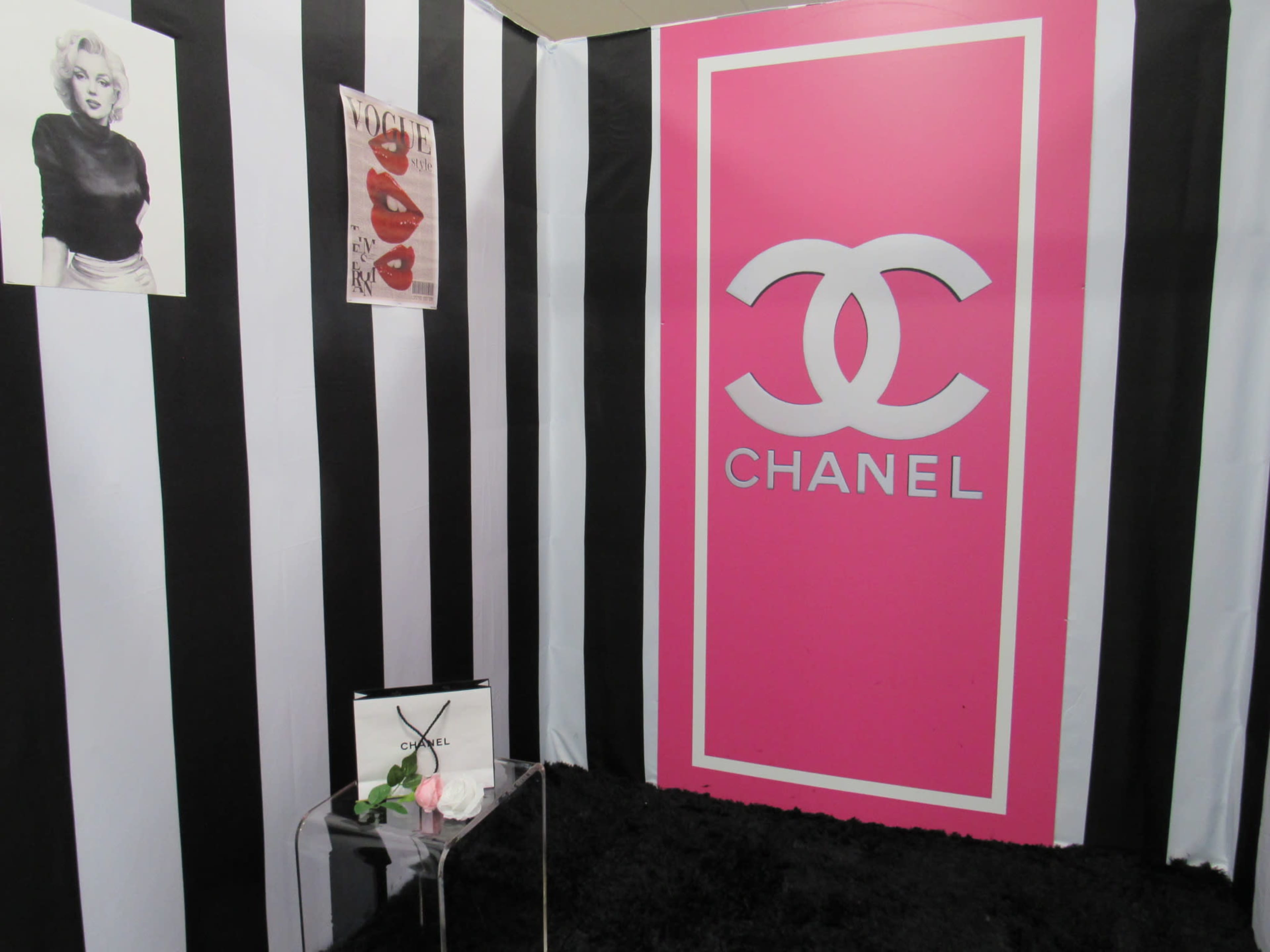 The image shows a corner set with black and white striped walls featuring a large pink Chanel logo and decorative elements, including a poster and a clear display stand with a flower and a magazine.