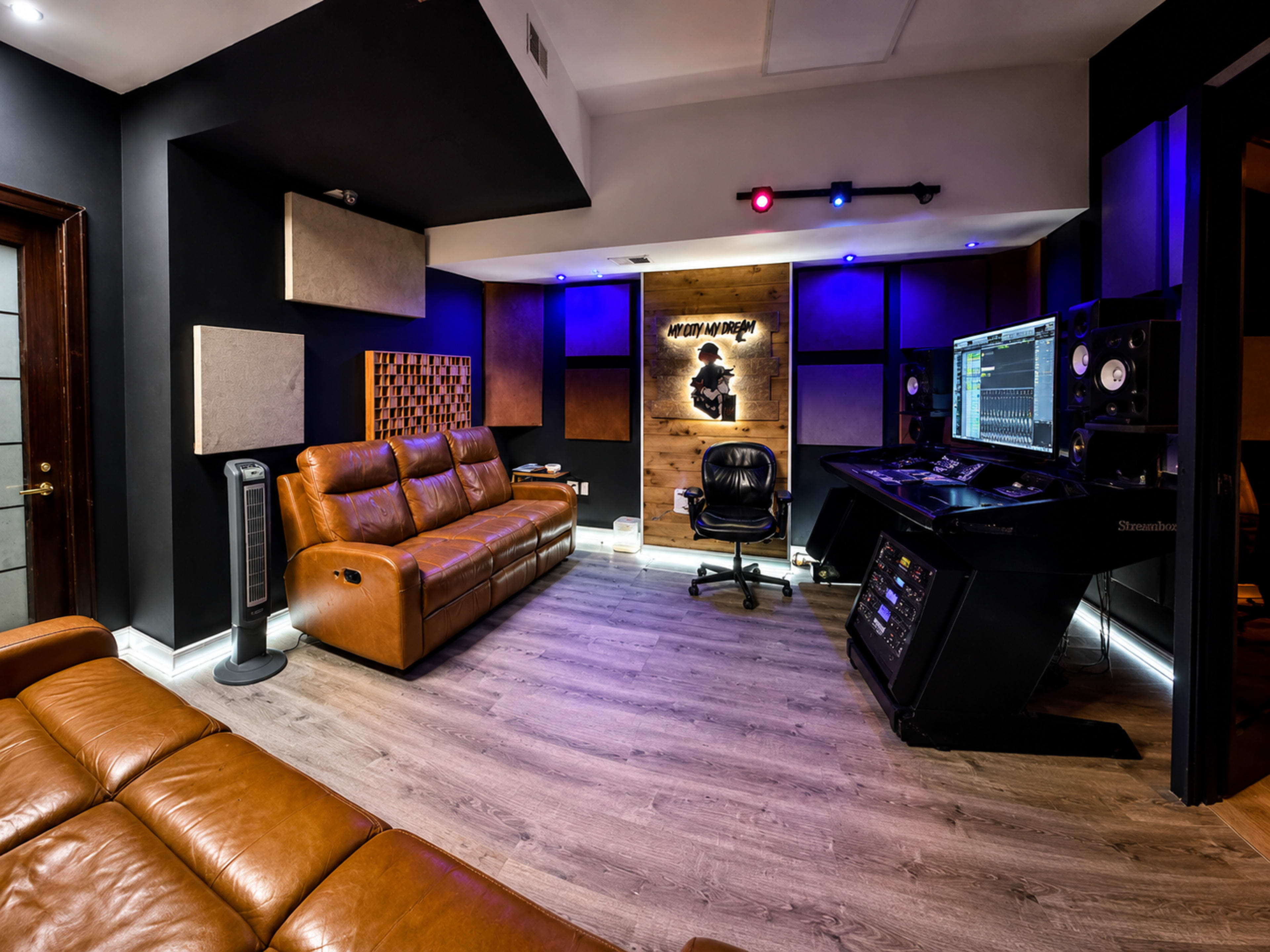 The image shows a modern recording studio featuring a leather sofa, a black swivel chair, acoustic panels on the walls, and a digital audio workstation setup with colorful lights.