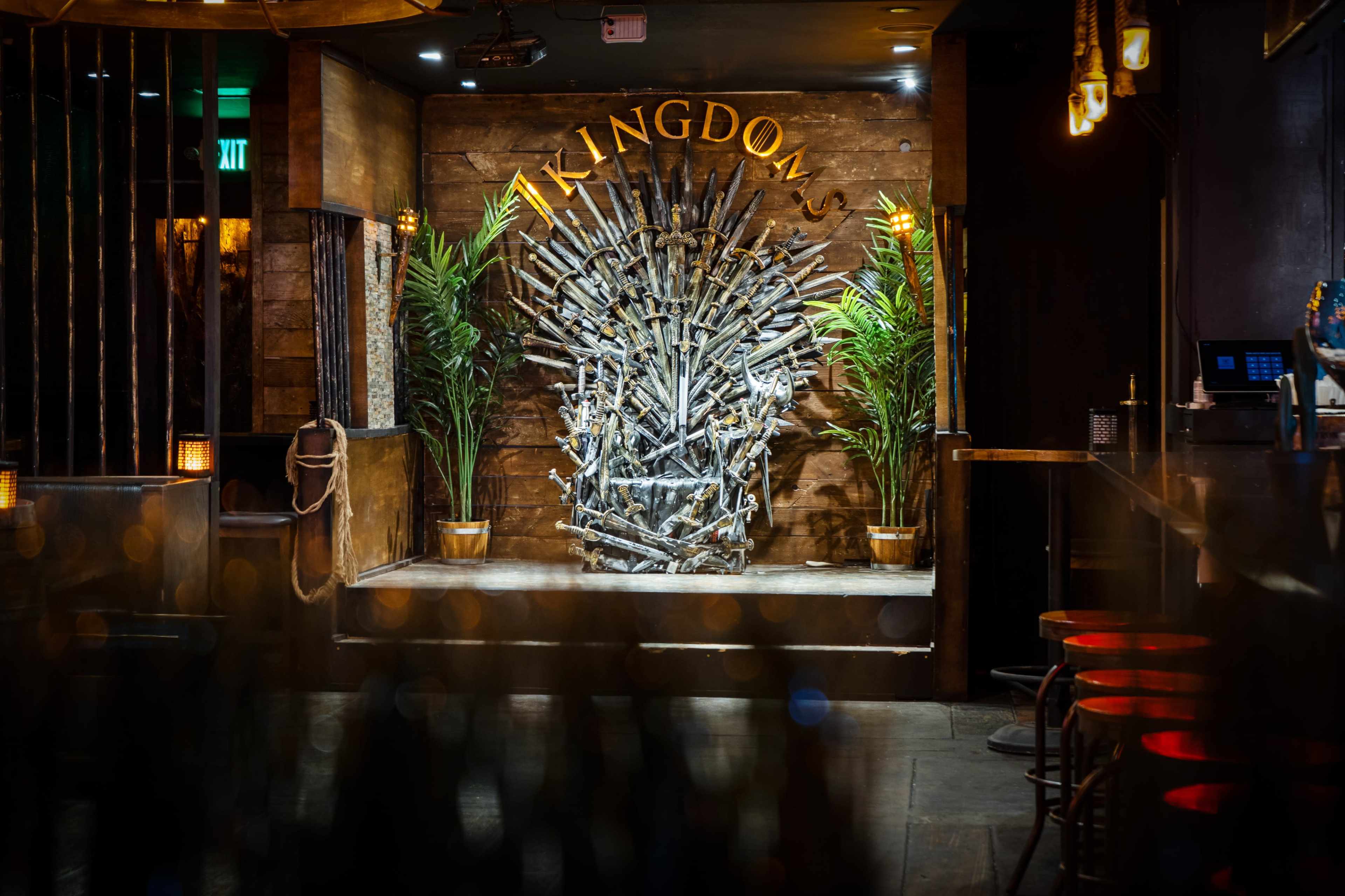 A themed bar with a prominent throne made of swords and a wooden wall that reads "KINGDOMS," surrounded by potted plants.
